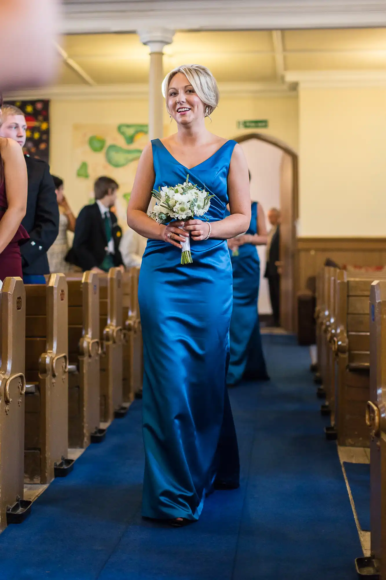 A woman in a blue dress holding a bouquet walks down a church aisle, smiling, with guests in the background.