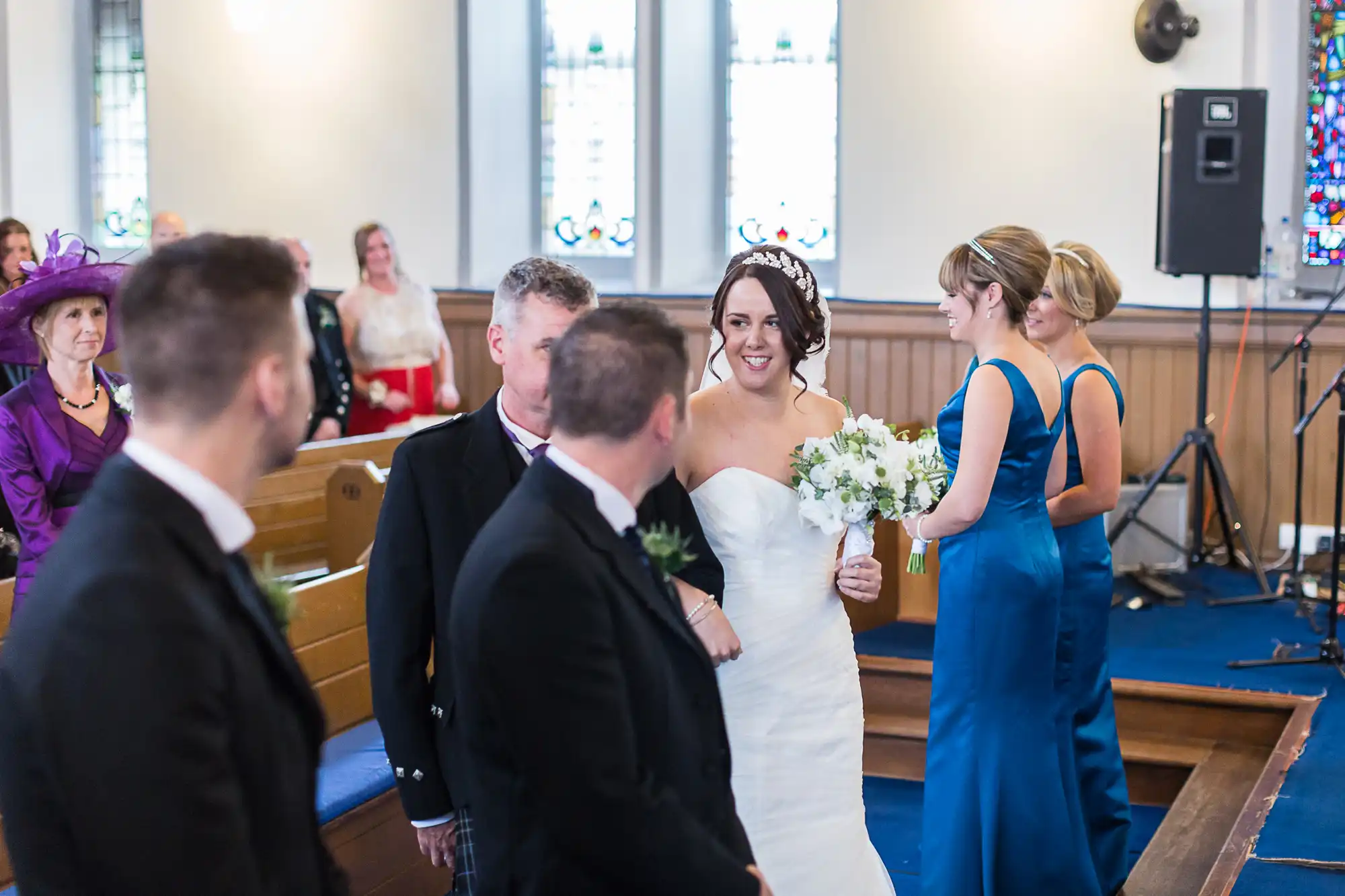 A bride smiling, holding a bouquet in a church, with wedding guests and a groom in the foreground. stained glass windows and a band setup are visible in the background.