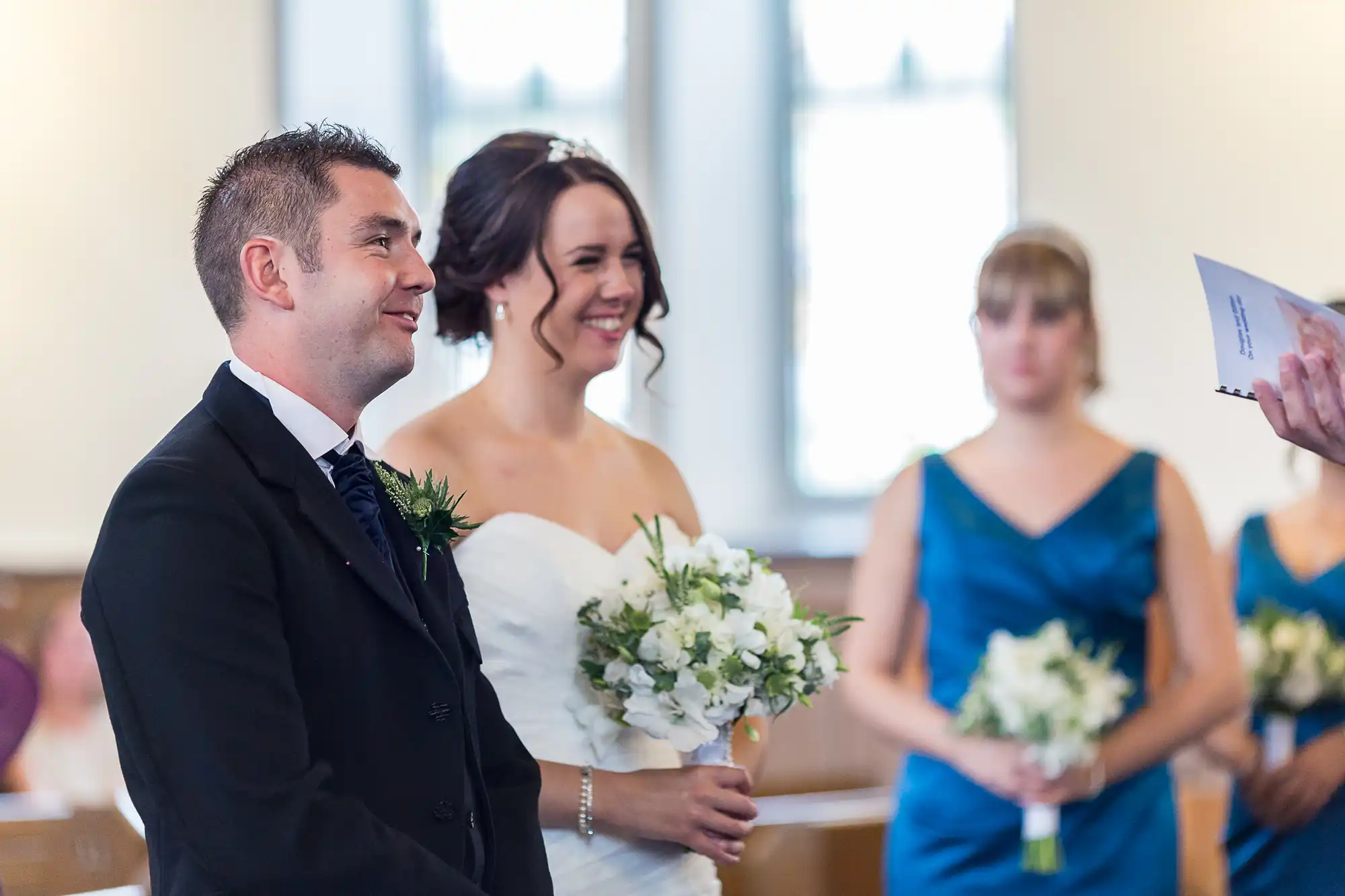 A bride and groom smiling during a wedding ceremony, holding hands, with bridesmaids in blue dresses in the background.