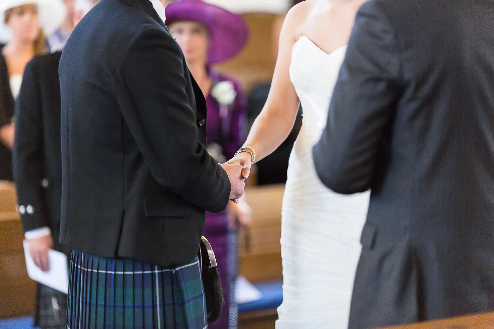 A bride and groom holding hands at their wedding ceremony, seen from behind a guest in a kilt and another in a purple hat.