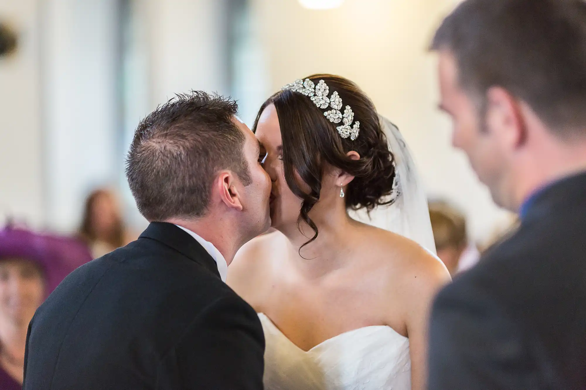 A bride and groom kissing during a wedding ceremony, with guests visible in the background.