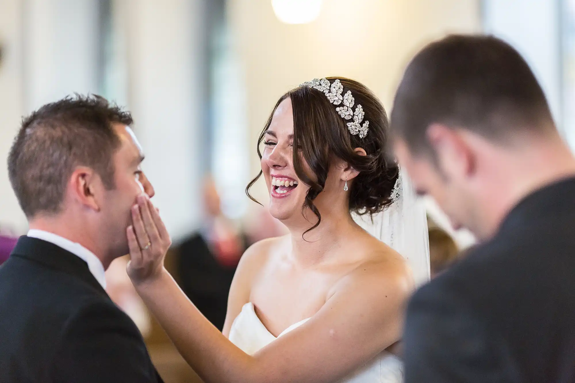 Bride laughing while touching a man's face, another man in foreground, indoors at a wedding reception.