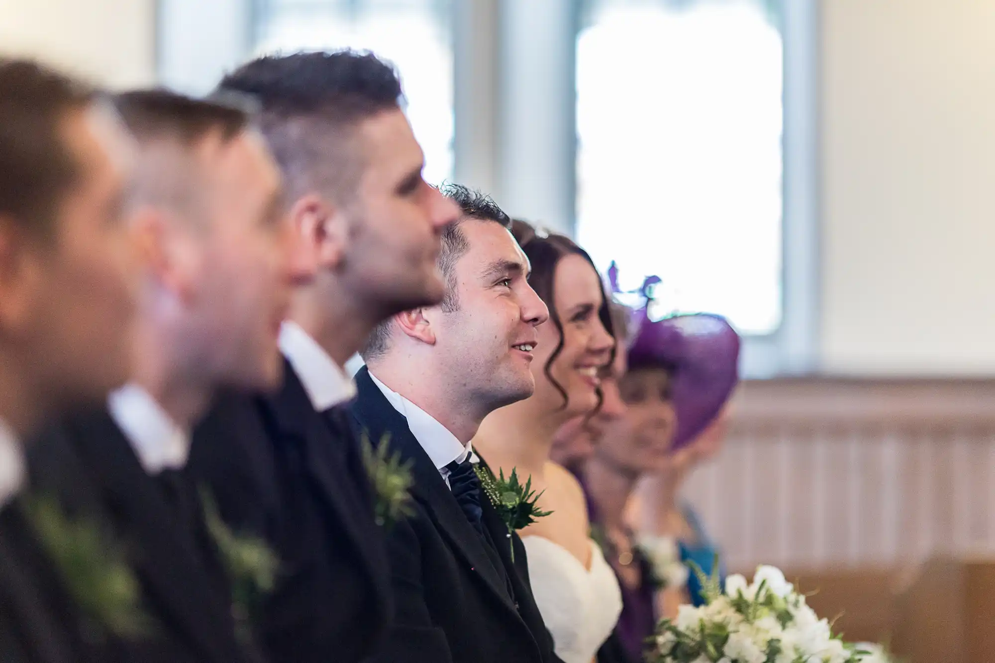 Guests in formal attire, smiling and focused on an event off-camera in a church setting.
