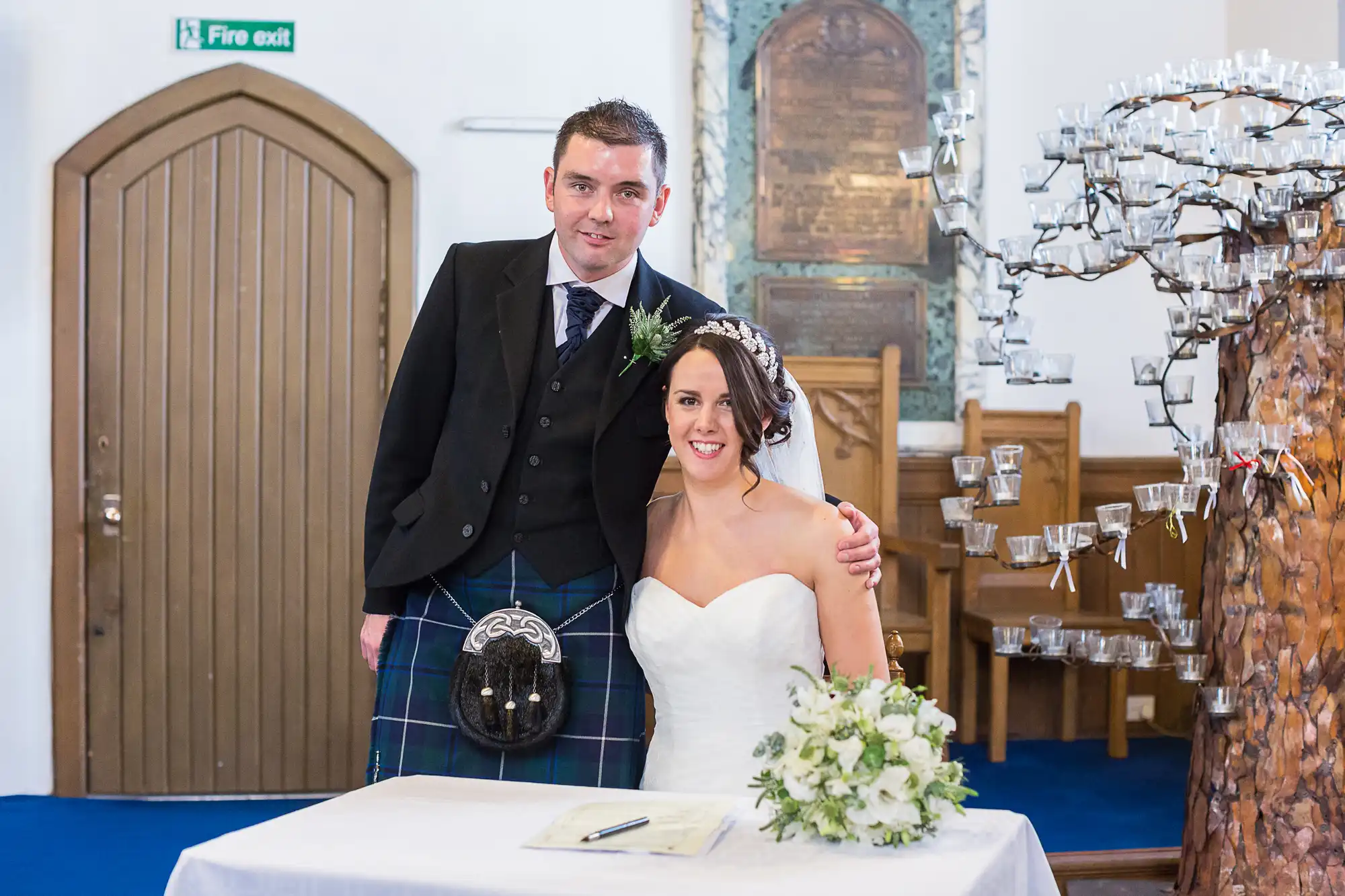 A bride and groom smiling at the camera in a church, the groom in a kilt and the bride holding a bouquet.