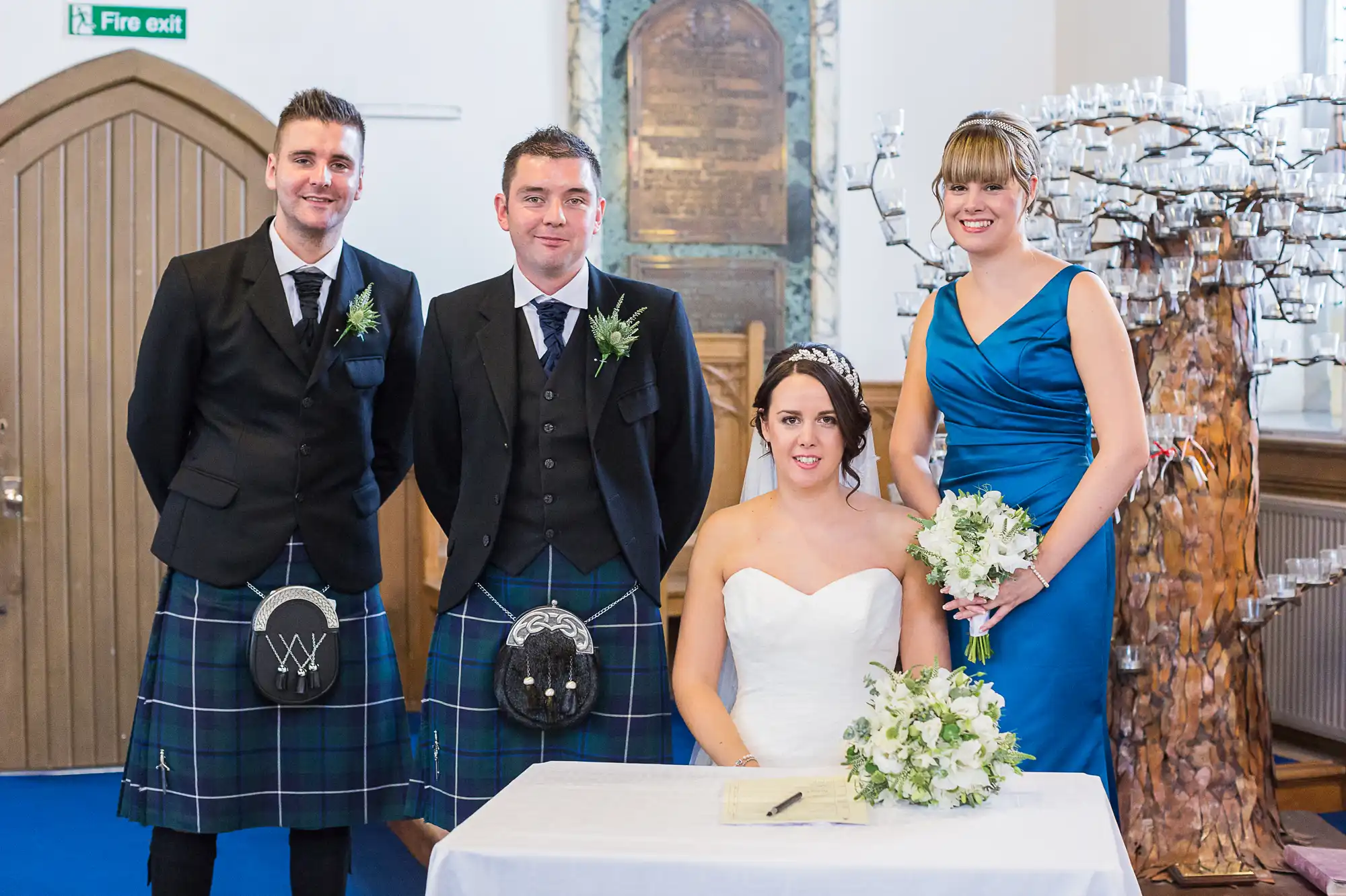 Four people dressed for a wedding posing in a church, including two men in kilts and a bride and bridesmaid holding bouquets.