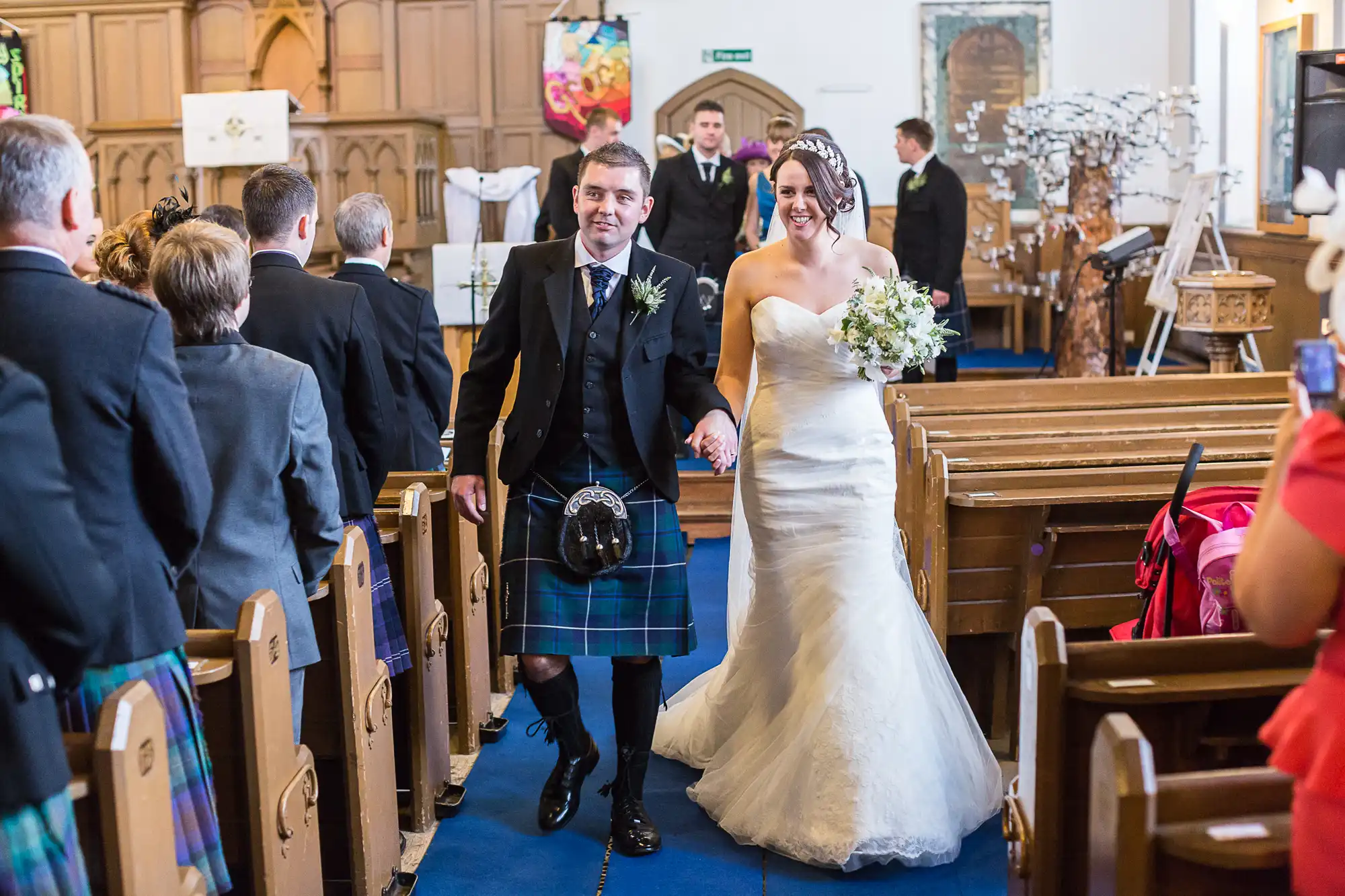 A bride in a white dress and a groom in a kilt walk down the aisle of a church, smiling, as guests look on.