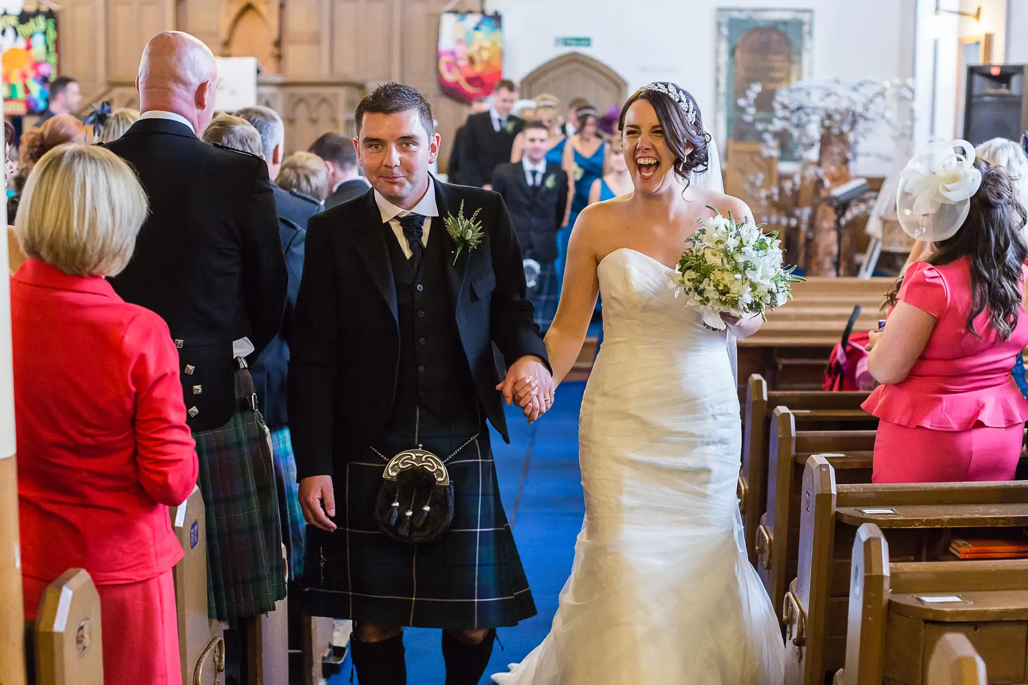 A bride and groom holding hands walk down the aisle, smiling, surrounded by guests in a church. the groom wears a kilt, and the bride is in a white gown.