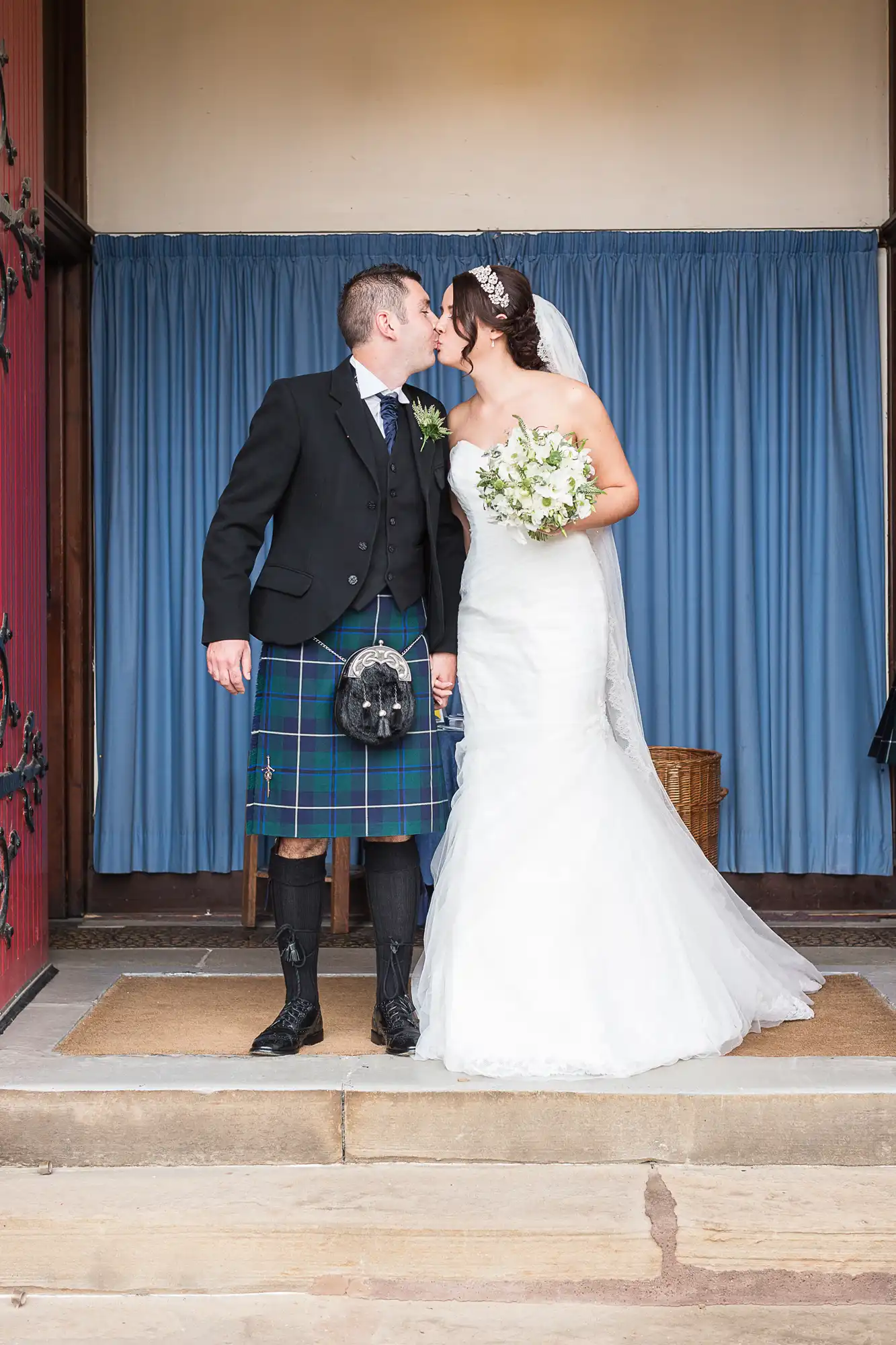A bride in a white dress and a groom in a kilt and sporran share a kiss at a doorway with blue curtains.