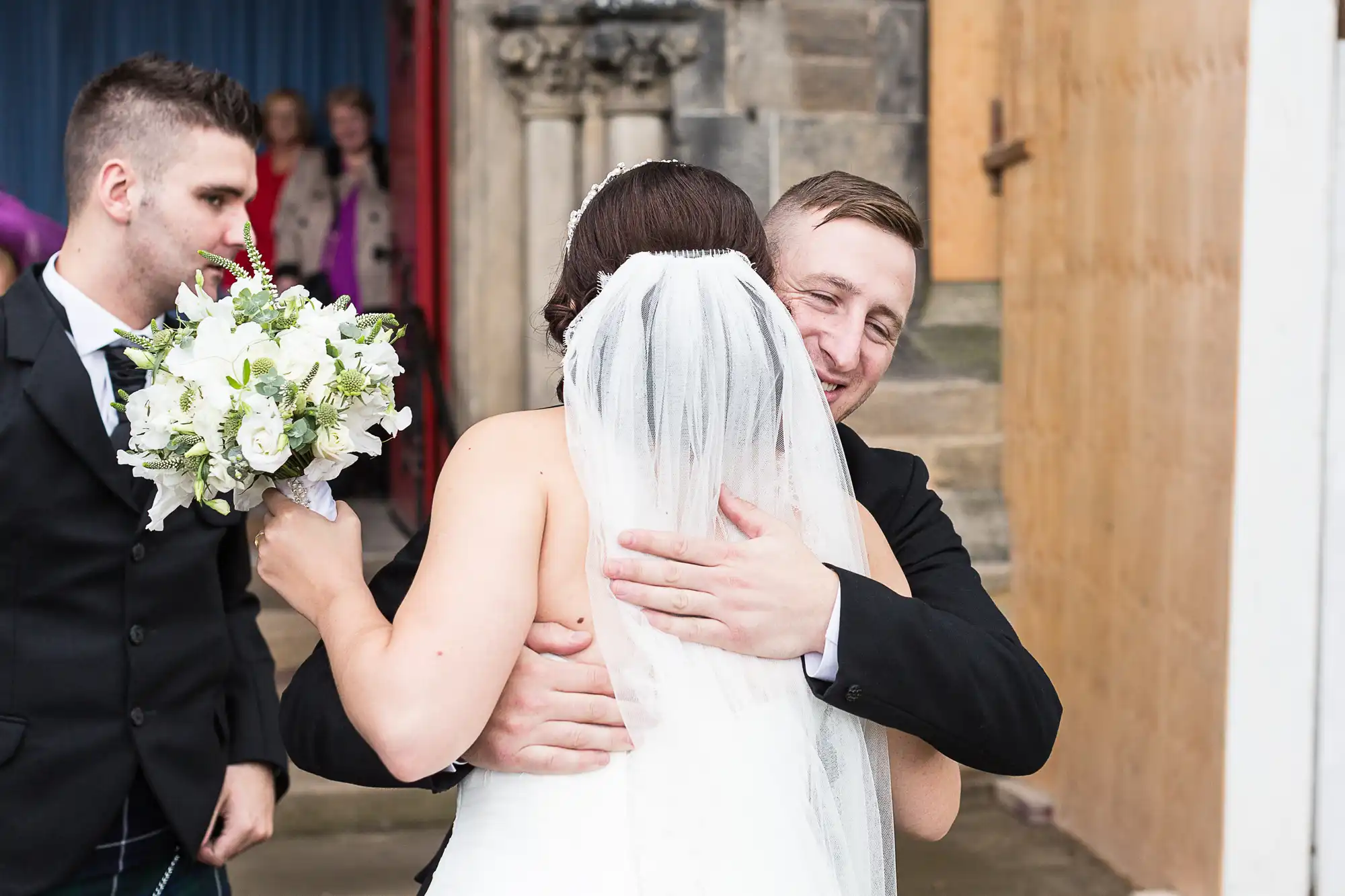 A bride and groom embracing outside a church, the groom smiling joyfully, with guests in the background.