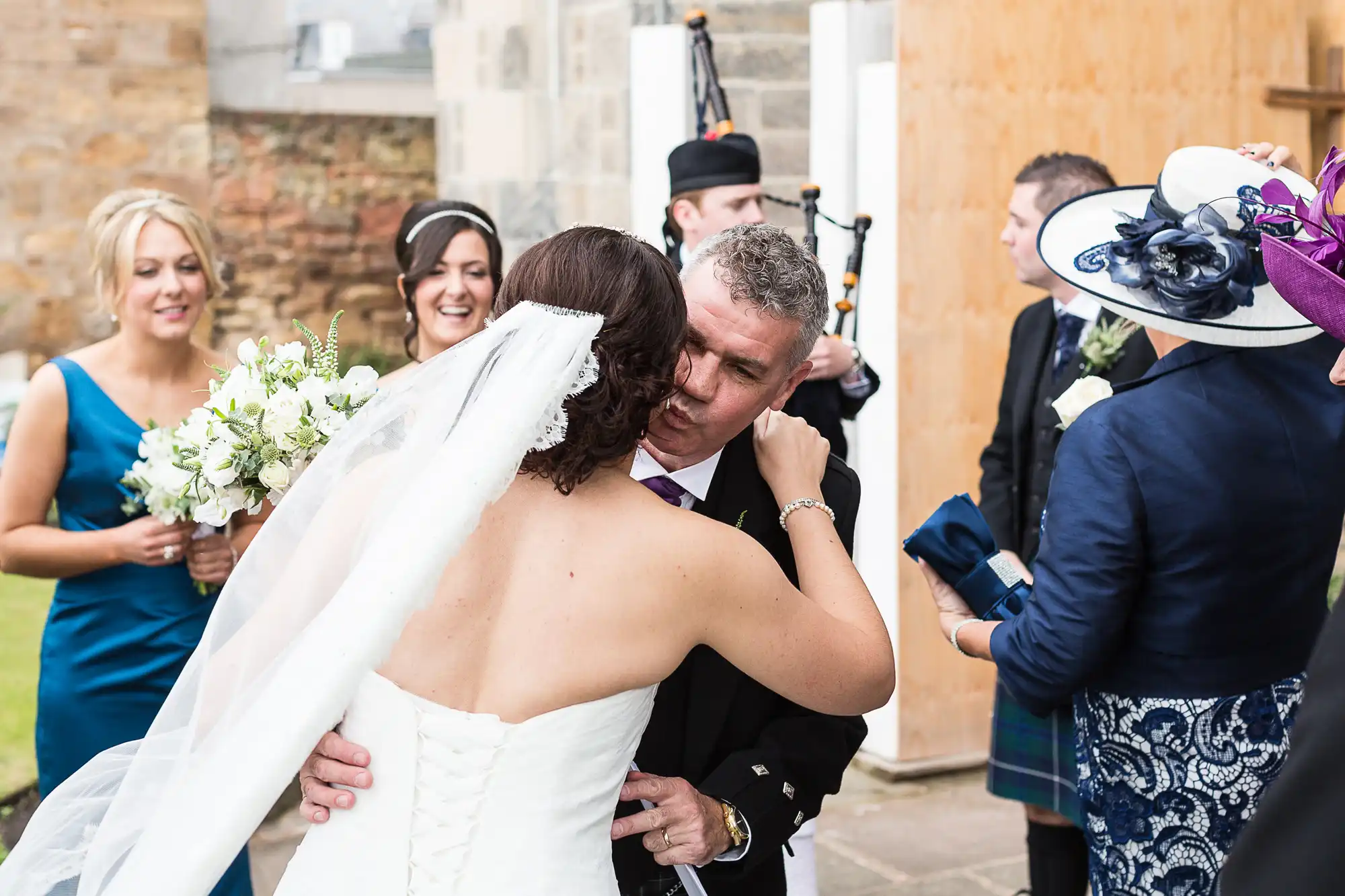 A bride in a white dress embracing a man in a suit at a wedding ceremony, with guests and a bagpiper nearby.