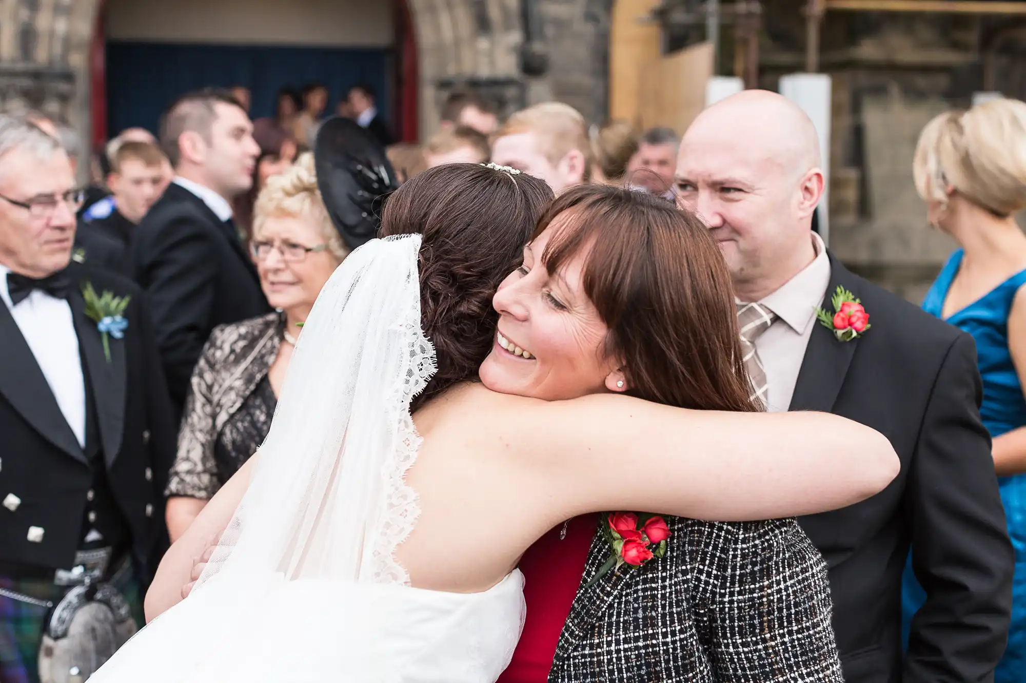 A bride in a white dress hugging a woman in a black dress at a wedding, with guests in the background, including a man in a kilt.