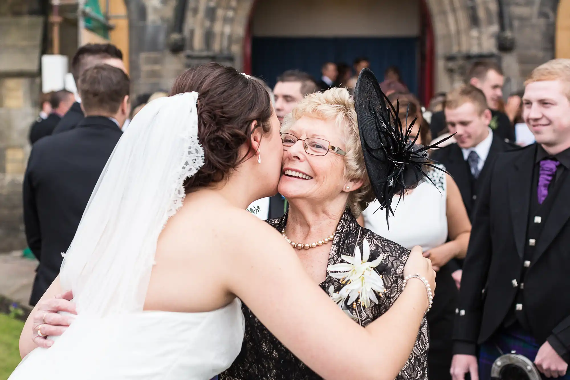 A bride in a white dress kisses an elderly woman wearing a black hat and floral dress outside a church, surrounded by wedding guests.