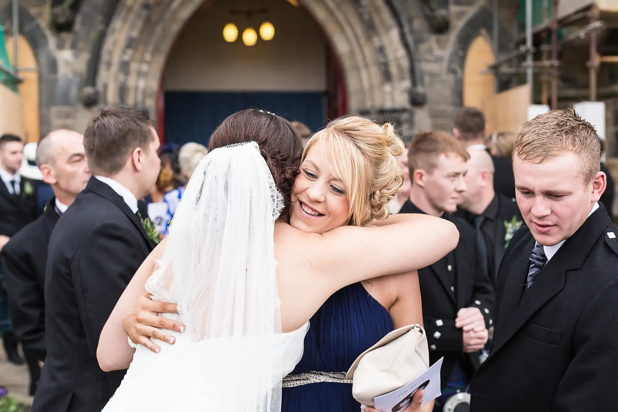 A bride and a woman in a blue dress embrace outside a church, surrounded by guests.