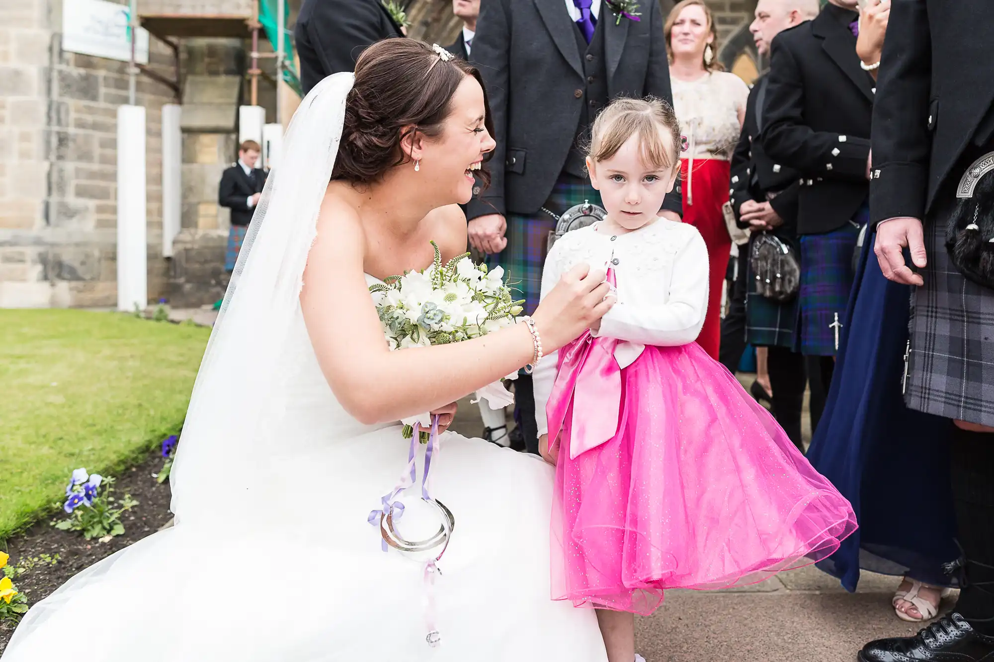 Bride in white dress holding bouquet interacting with a young girl in a pink dress outside a church, with guests in the background.