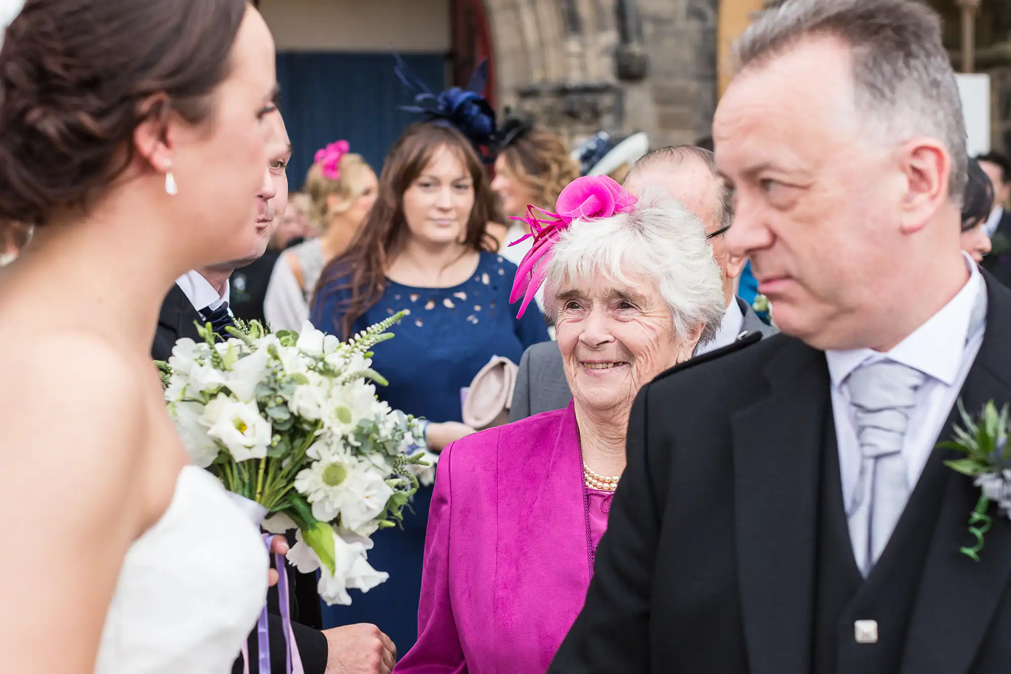 Bride holding a bouquet converses with an elderly woman in a pink hat and a man in a suit, with other guests in the background.