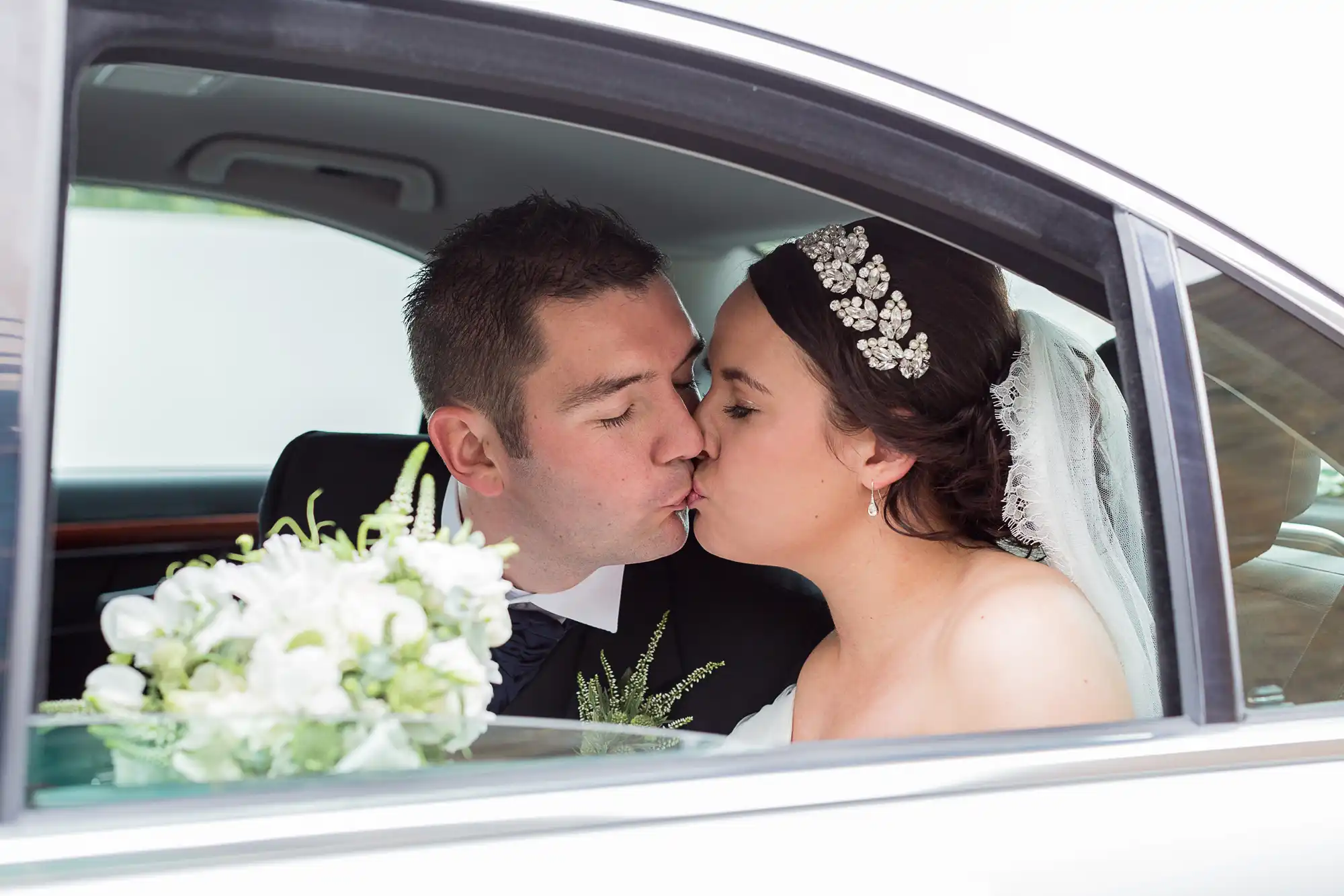 A newlywed couple kissing inside a car, with the bride holding a bouquet and wearing a tiara and veil.