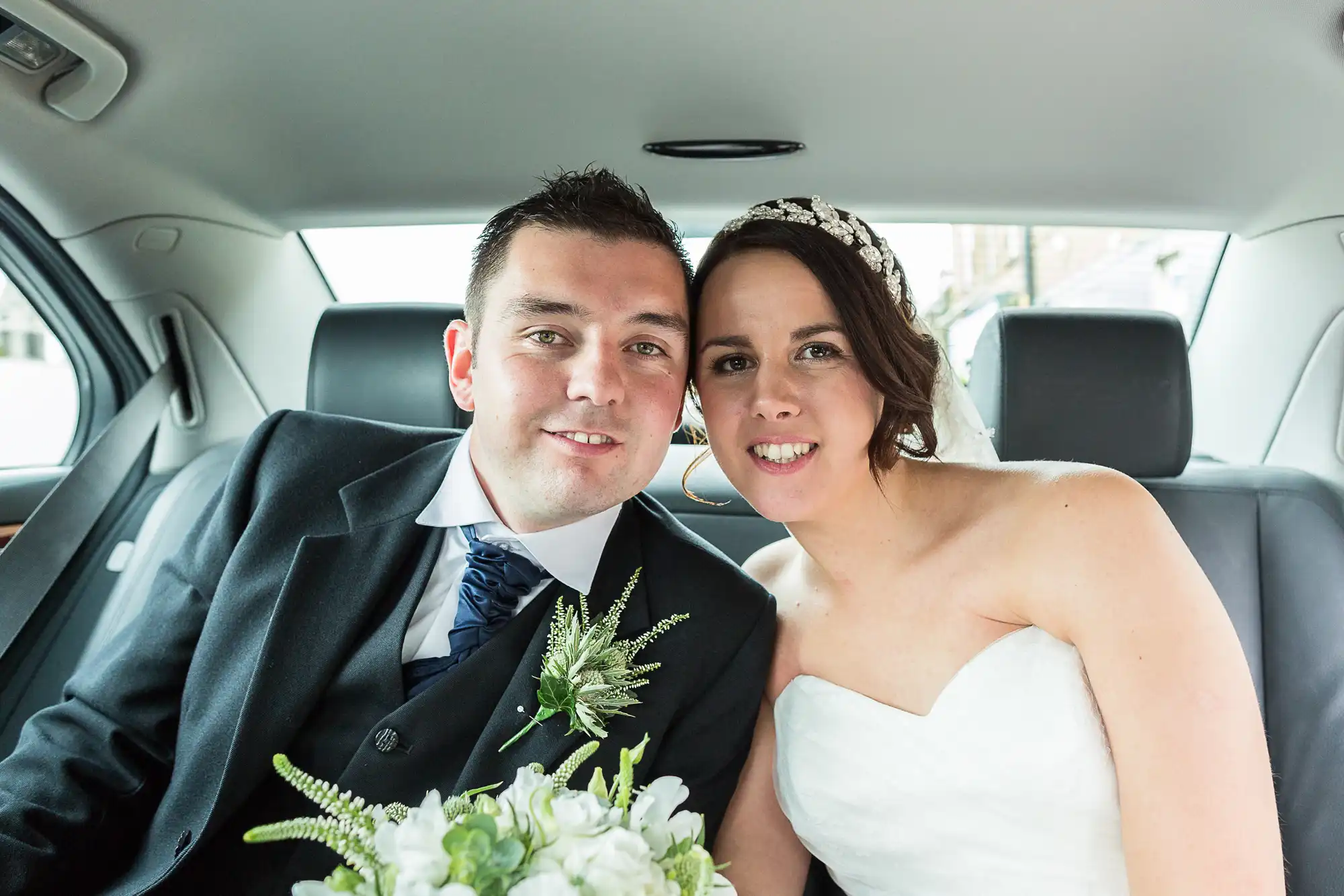 A newlywed couple, a man and a woman, smiling inside a car. the man wears a black suit and the woman a white wedding dress.