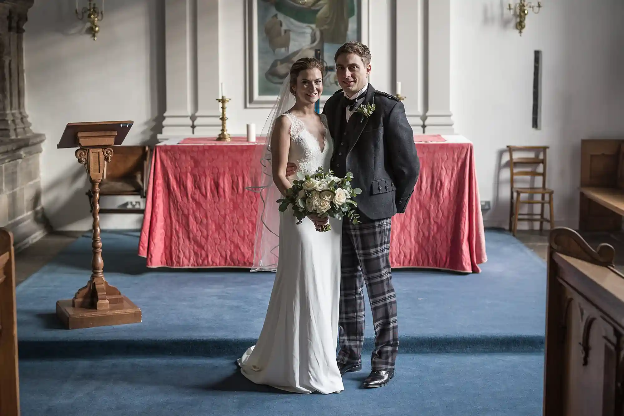 A bride in a white dress and a groom in a kilt and jacket stand together in a church, holding hands and smiling.