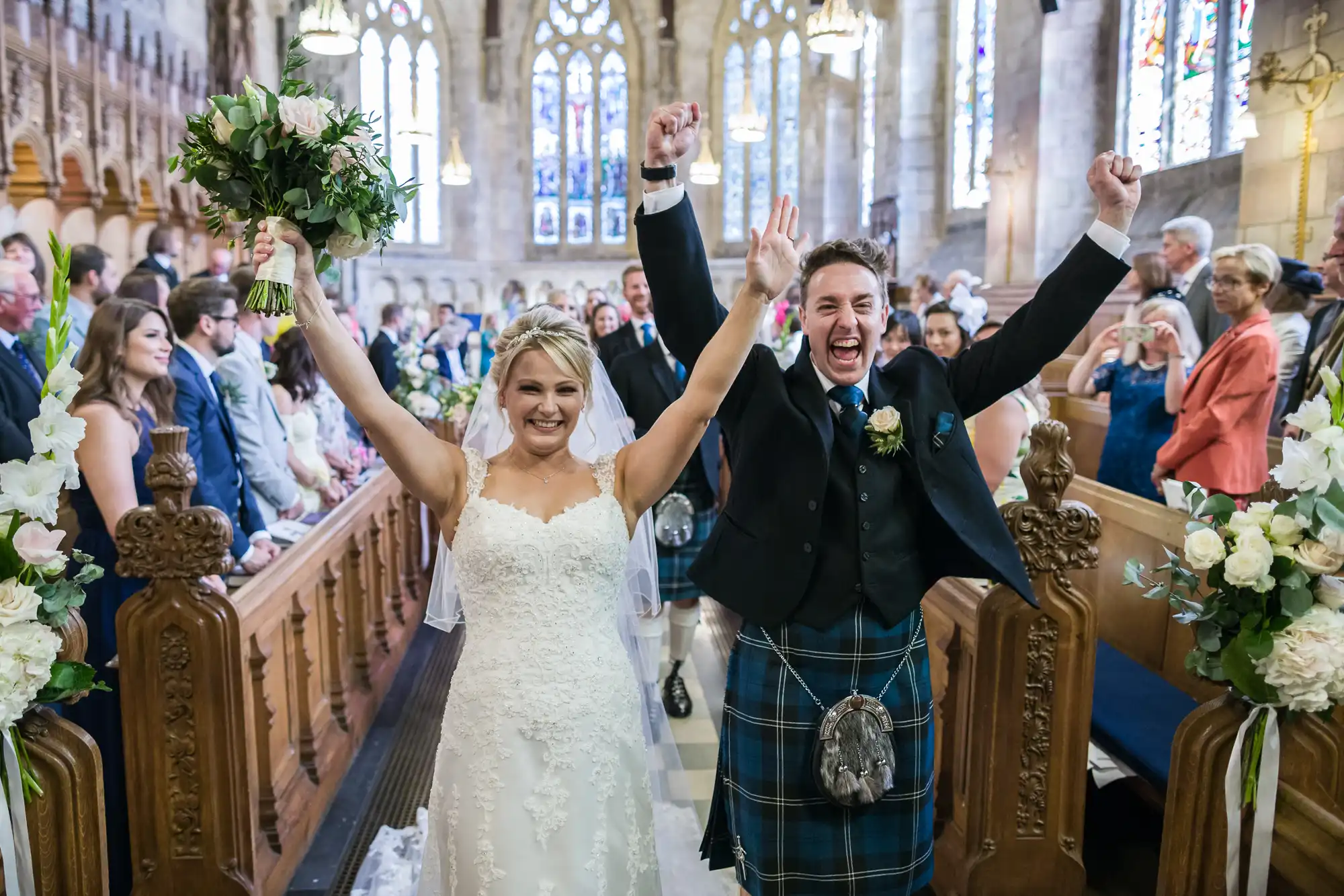 A bride and groom joyfully raise their arms, walking down the aisle of a packed church, as guests look on and cheer. The groom is dressed in a traditional Scottish kilt.