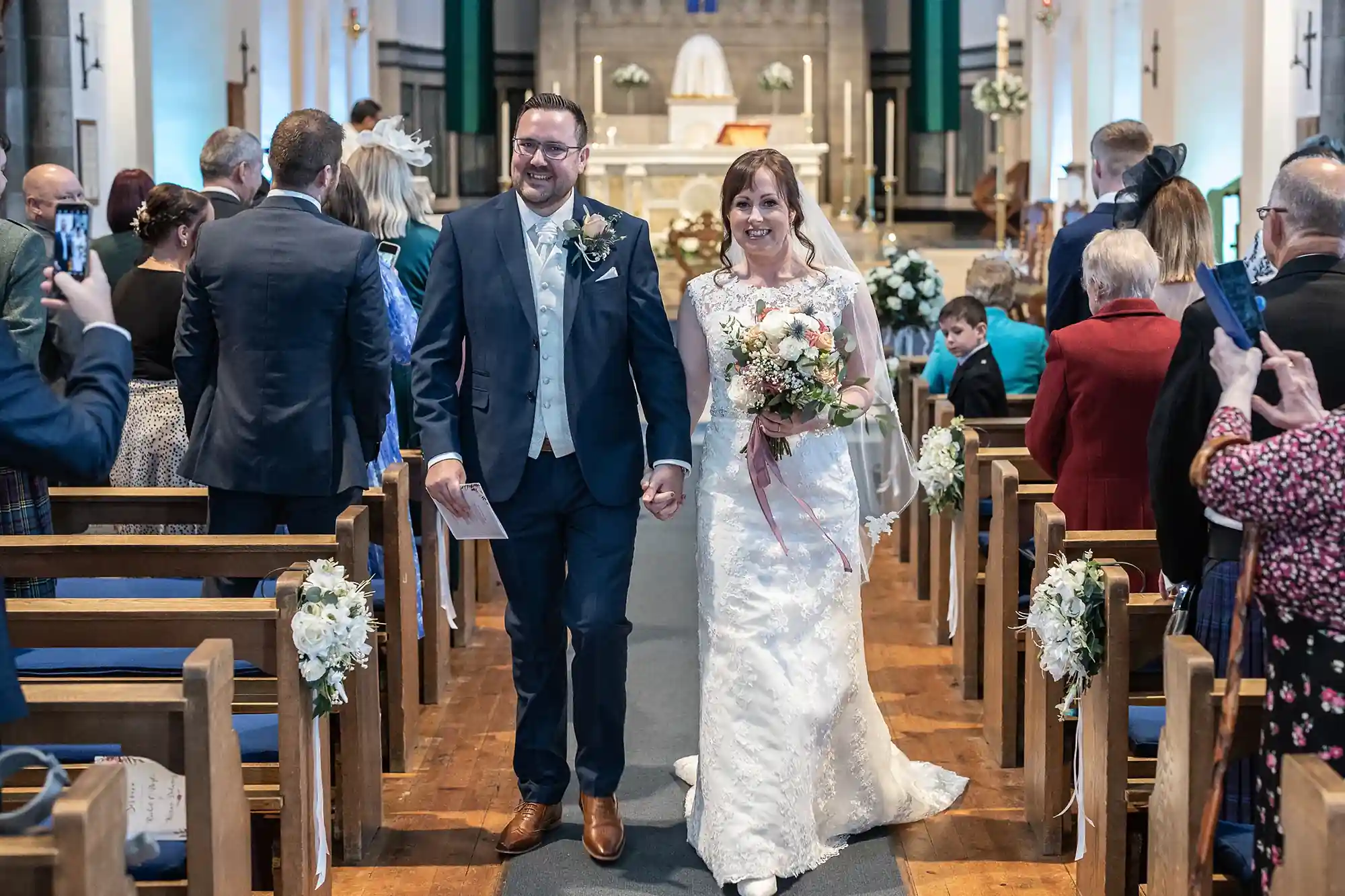 A bride and groom walk down the aisle of a church, smiling, as guests capture the moment with their smartphones.