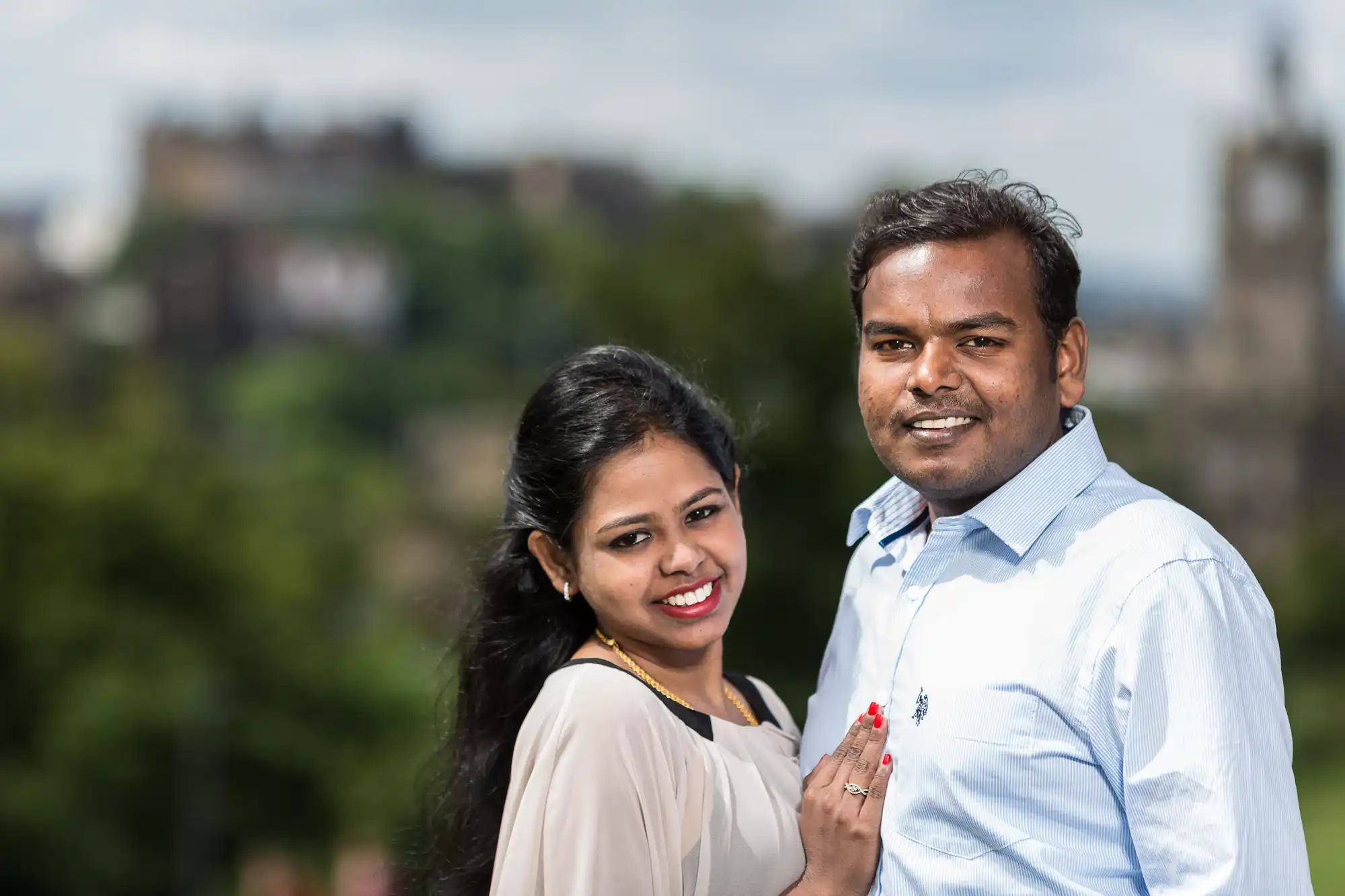 A smiling couple stands together outdoors with blurred greenery and historic buildings in the background.