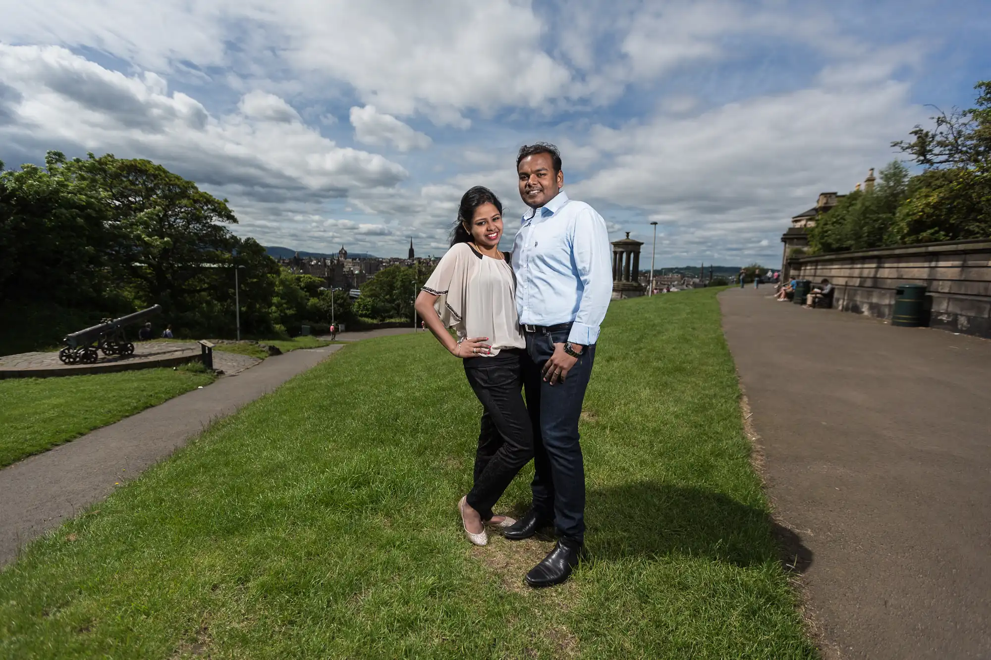 A woman and a man stand on a grassy area with a partly cloudy sky in the background. They pose side by side, smiling at the camera, with pathways and buildings visible around them.