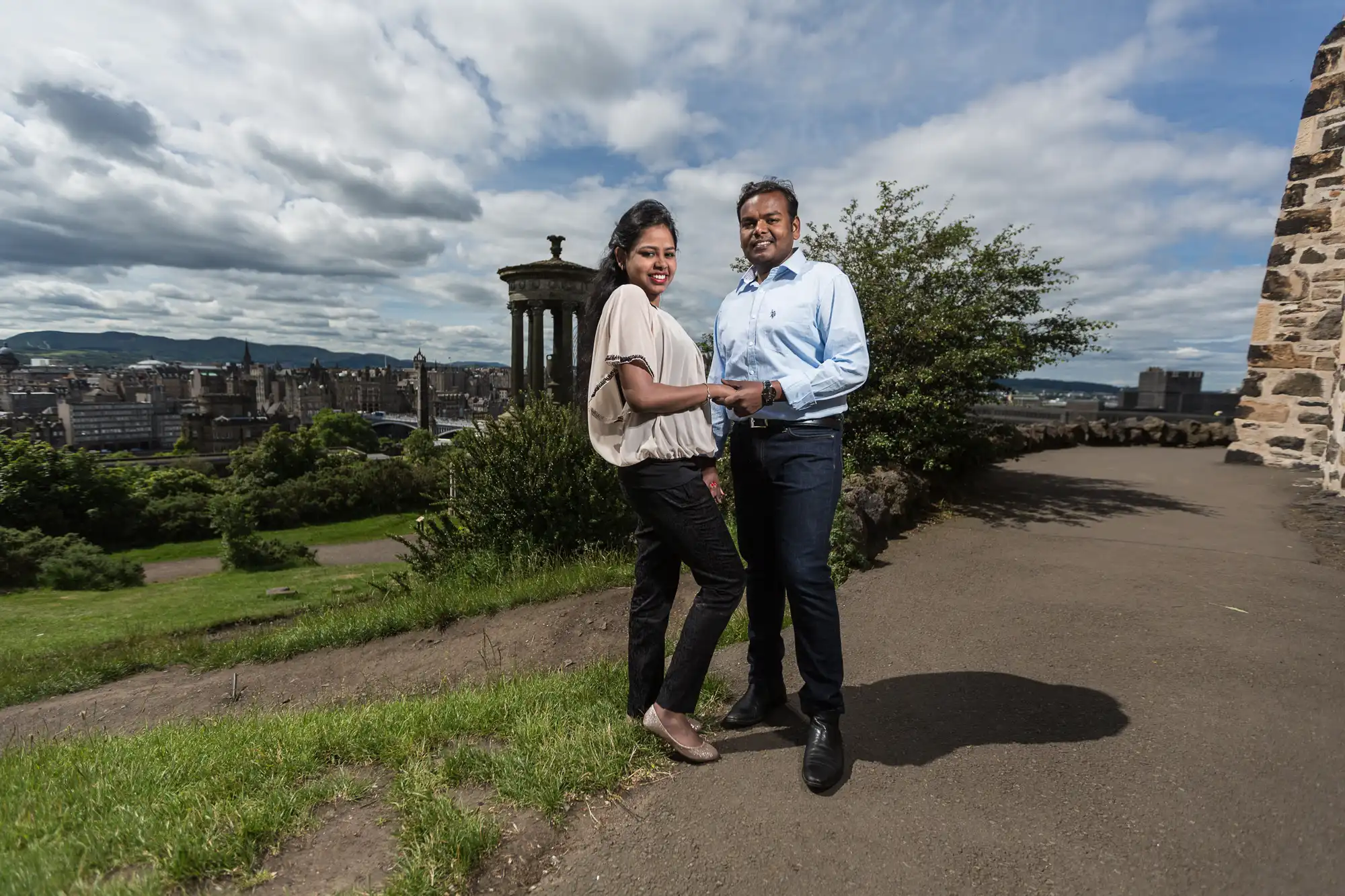 A couple stands on a walkway near a historic structure with a cityscape and cloudy sky in the background.