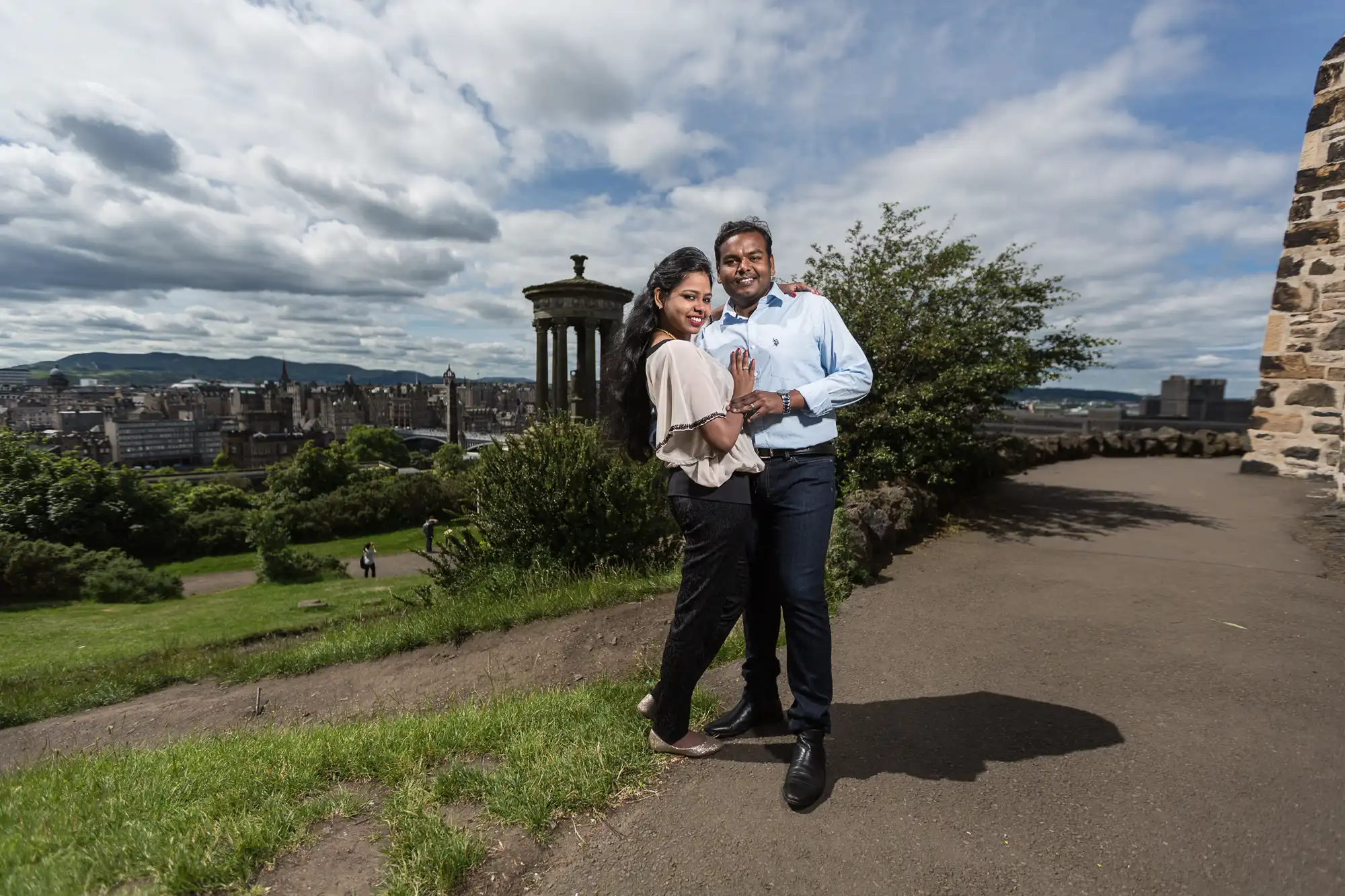 A couple stands together and smiles on a path with a historical cityscape and stone monument in the background on a partly cloudy day.