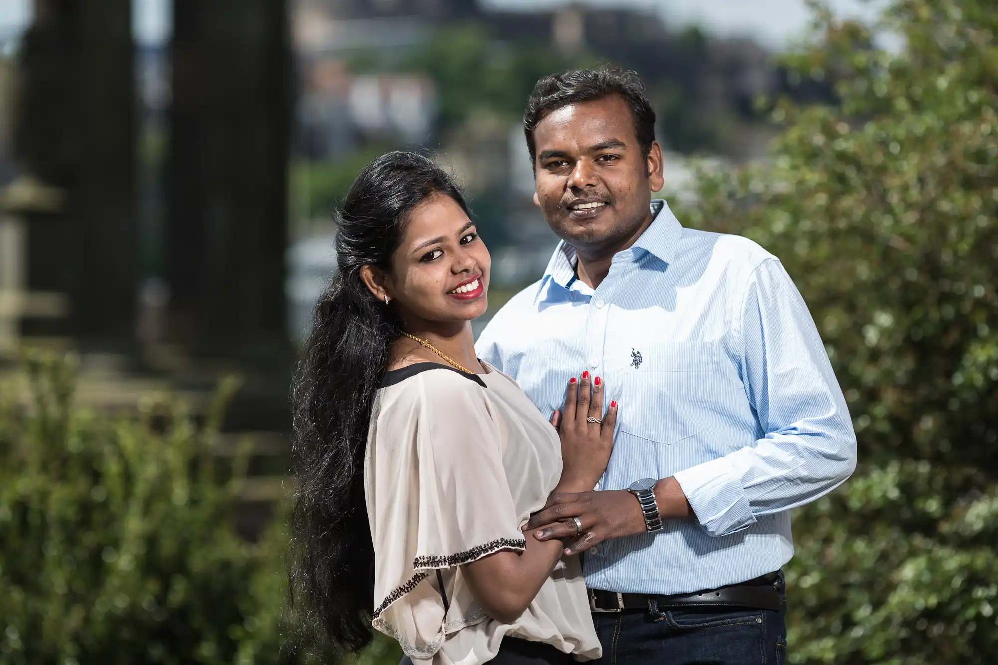 A couple poses together outdoors, smiling at the camera. The woman has long black hair and is wearing a light-colored blouse. The man has short black hair and is wearing a light blue shirt.