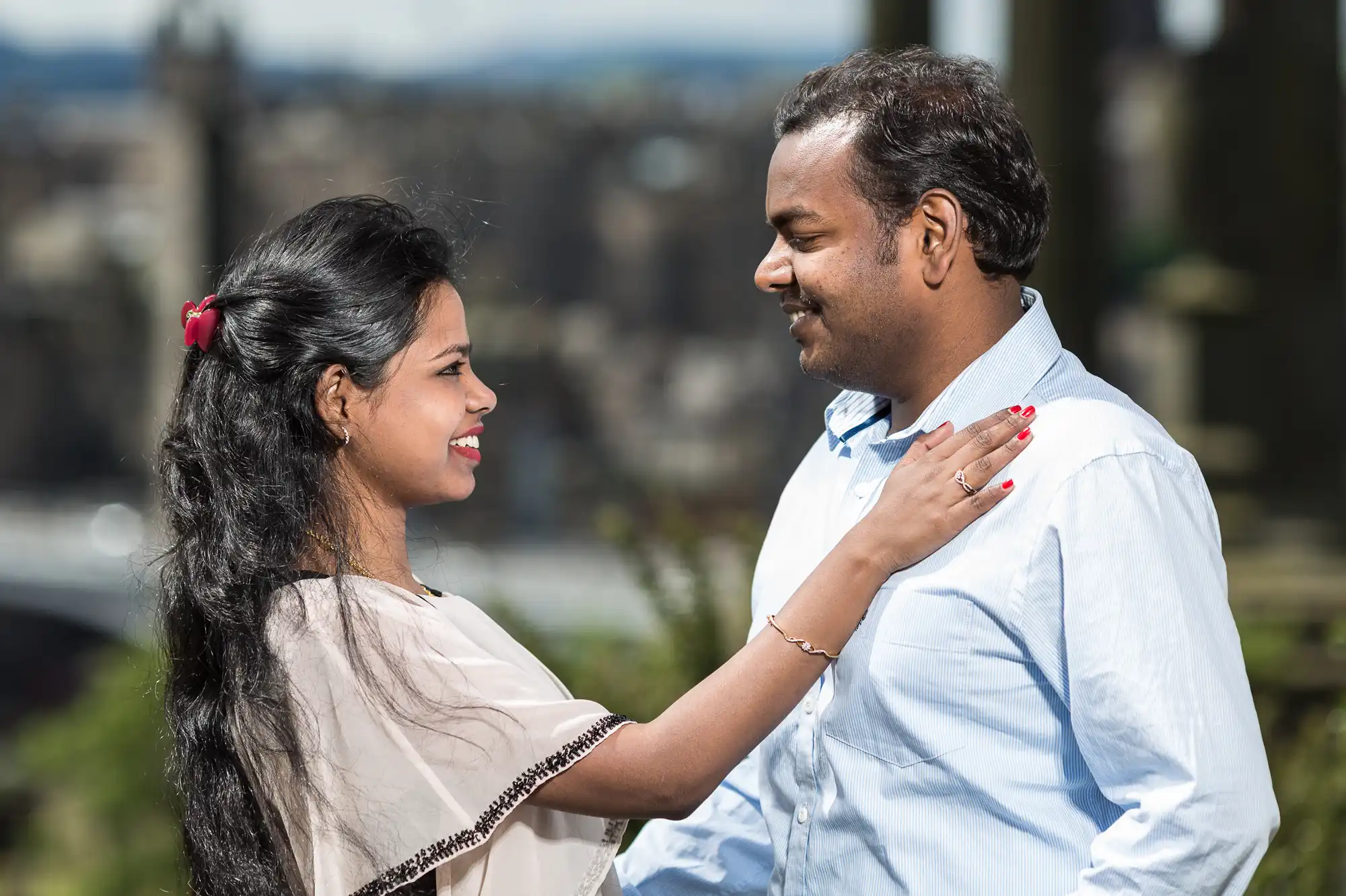 A woman and a man looking at each other and smiling outdoors, with the woman's hand resting on the man's shoulder. The background is blurred.