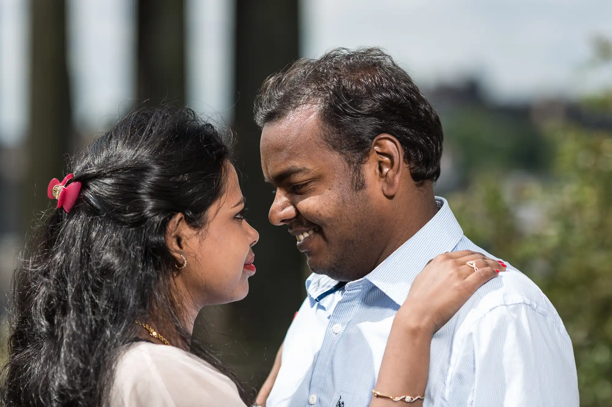 A couple stands close, facing each other and smiling. The woman has her arms on the man's shoulders. They are outdoors with greenery in the background.