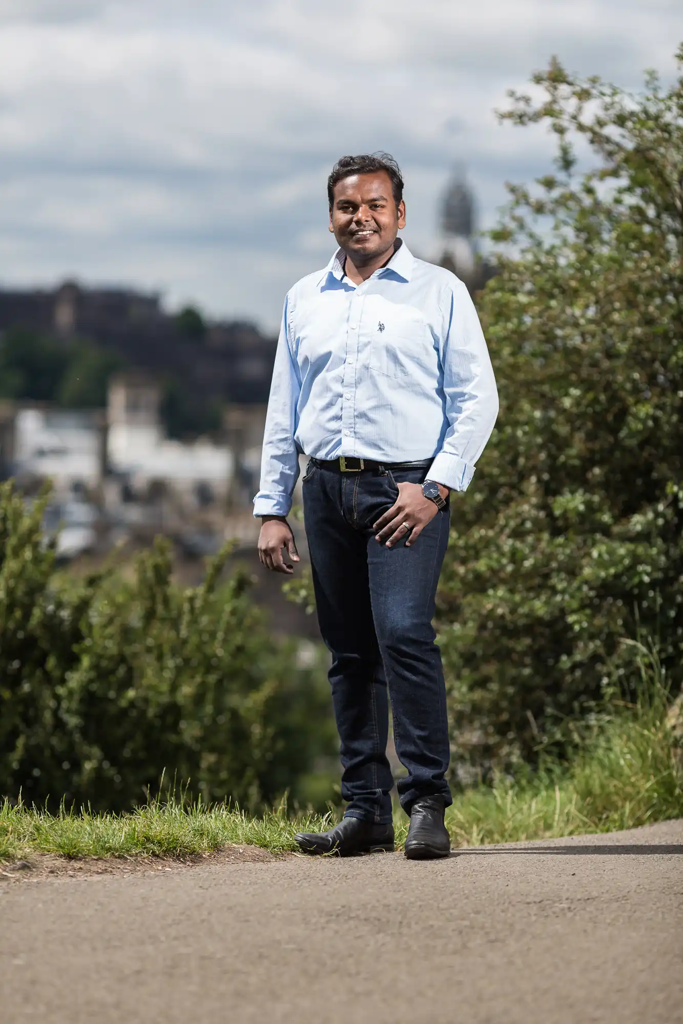 A man stands on a pathway outdoors, wearing a light blue shirt and dark jeans, with greenery and buildings in the background.