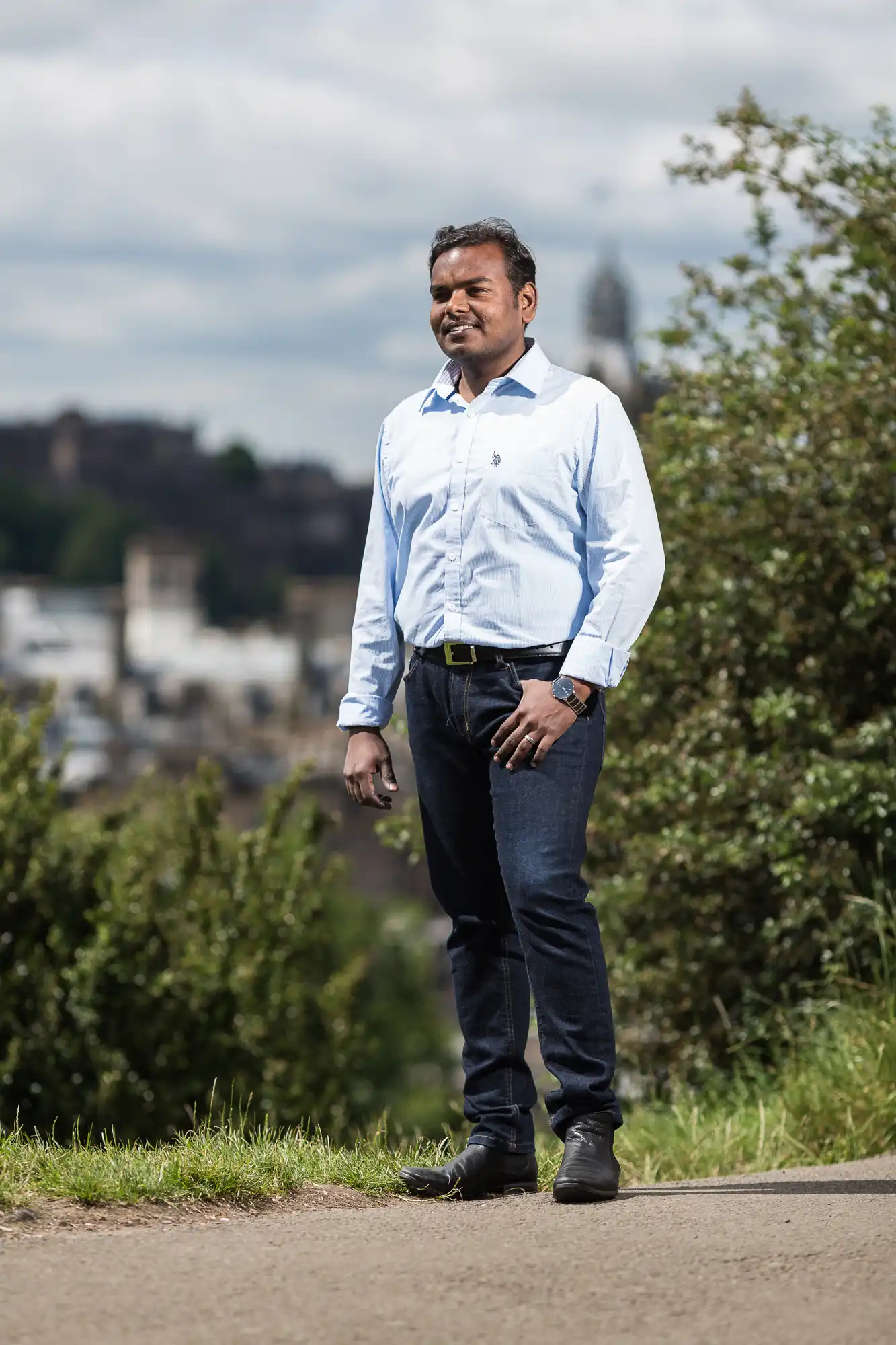 A man stands outdoors on a path, wearing a light blue shirt and dark jeans. Trees and buildings are visible in the background.