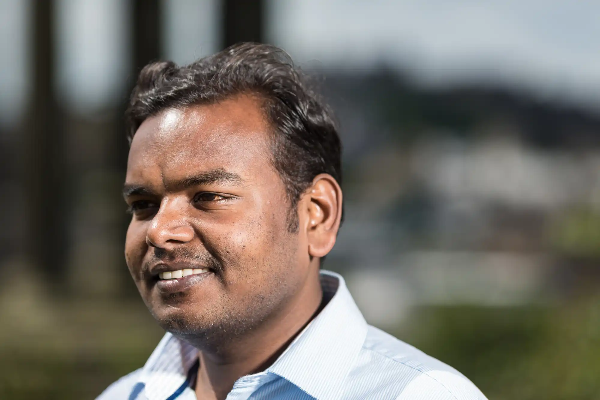 A man with short dark hair, wearing a light blue shirt, is smiling while standing outdoors with a blurred background of trees and buildings.