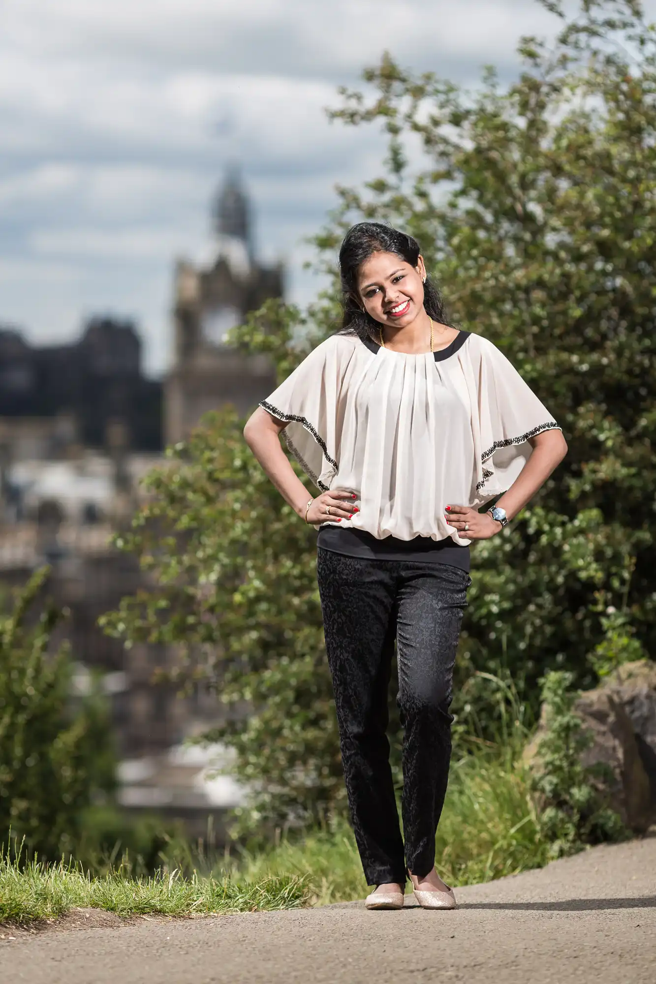 A woman stands outdoors on a path with greenery and a blurred cityscape in the background, smiling with her hands on her hips, wearing a white blouse and black pants.