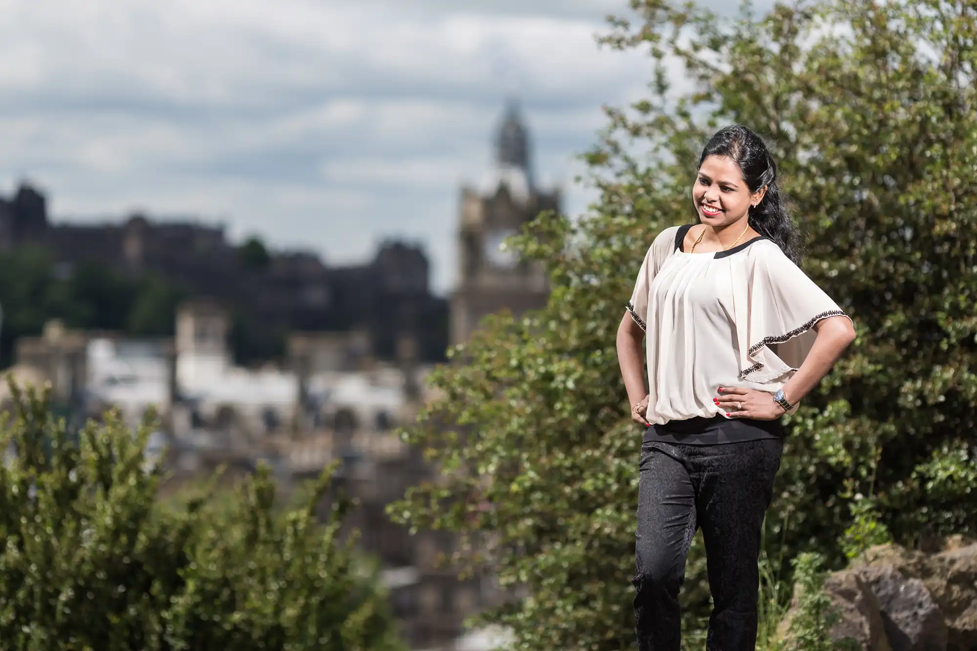 A woman stands outdoors in front of trees and a blurred cityscape. She is wearing a white top and black pants, with her hands on her hips.