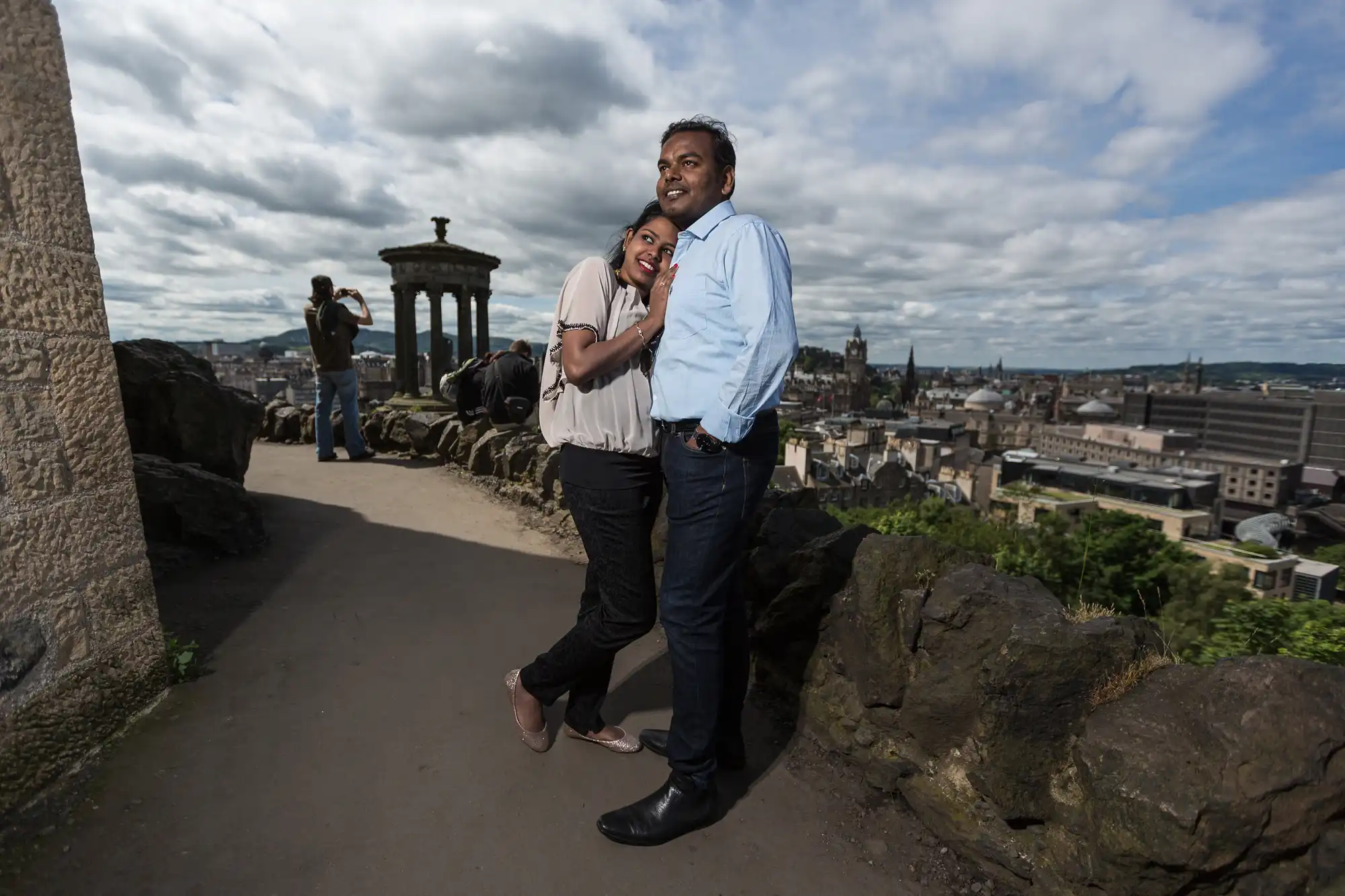 A couple poses together on a hilltop overlooking a cityscape. Another person is photographing the scenery in the background. The sky is partly cloudy.