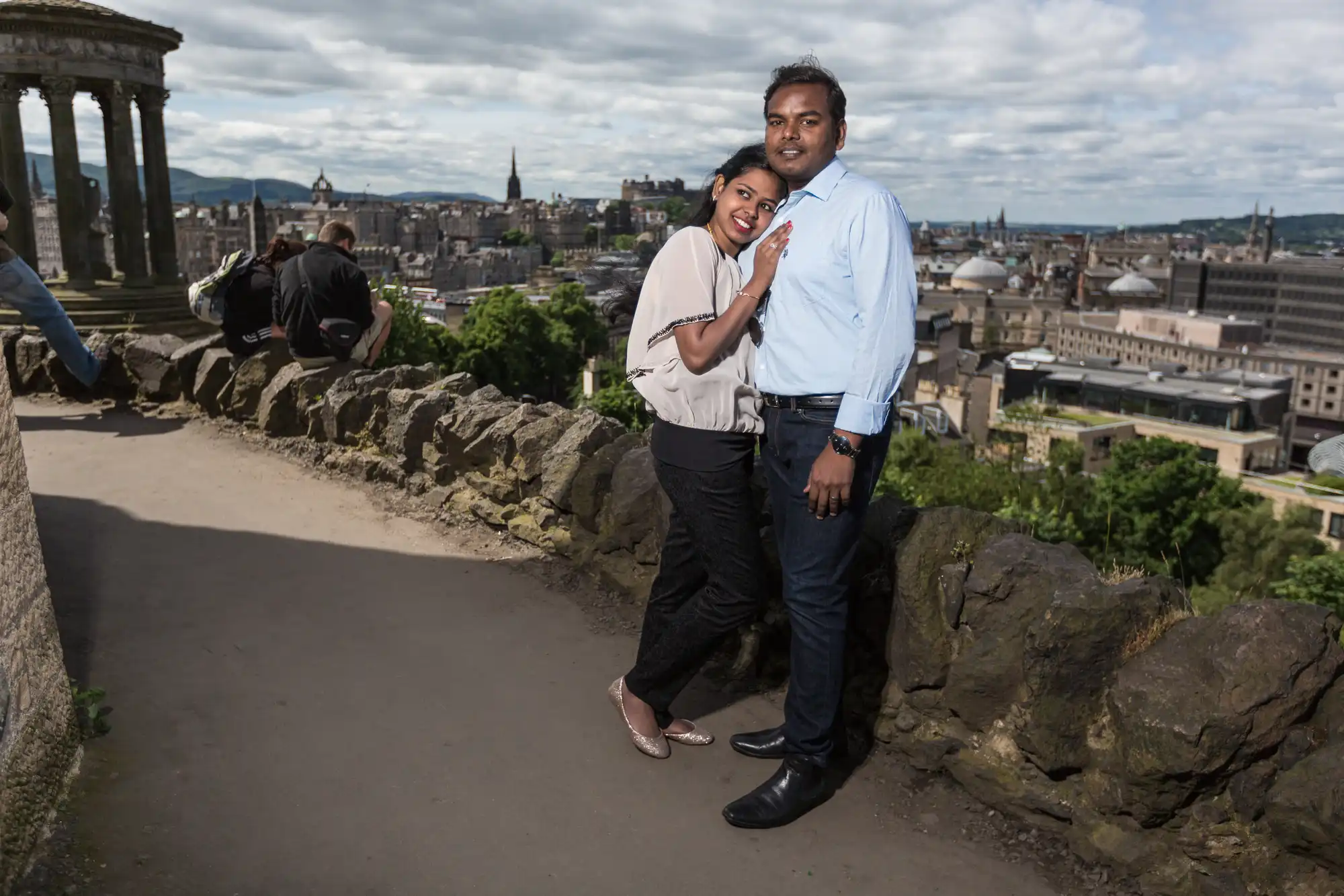 A couple stands together on an elevated path with a scenic city view in the background. The woman leans in, smiling, while the man stands next to her. The sky is partly cloudy.