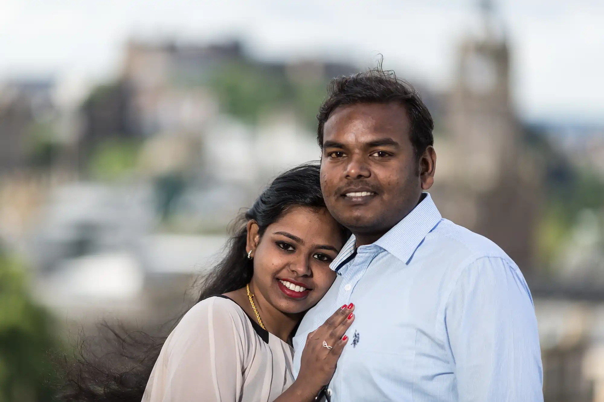 A couple stands together outdoors, the woman leaning her head on the man's shoulder, both smiling at the camera. A blurred landscape background with greenery and buildings is visible.