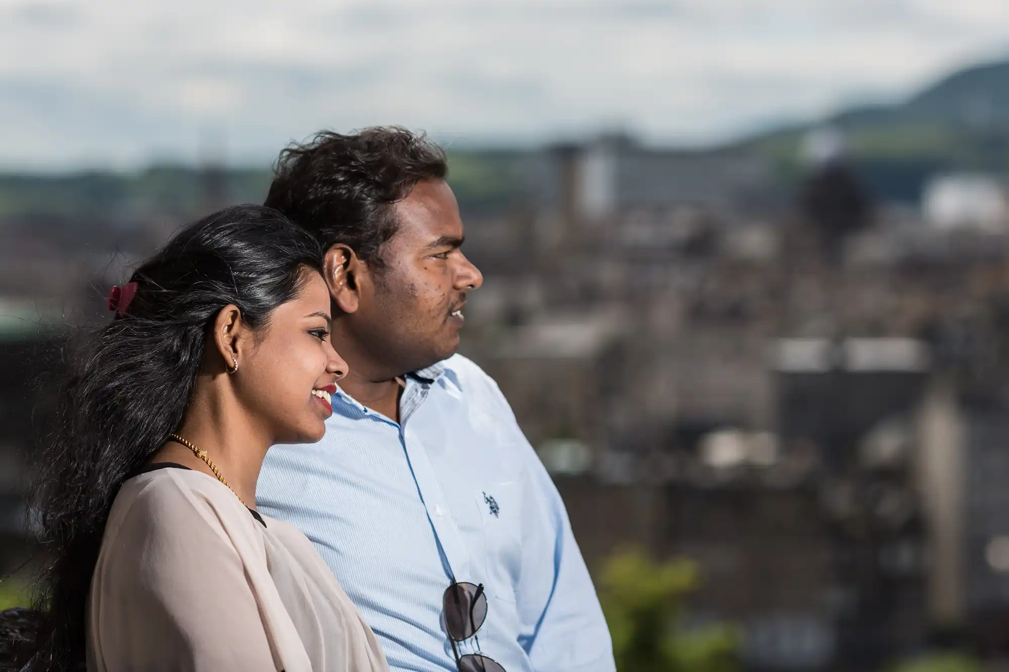 A man and a woman stand side by side, looking into the distance with a blurred cityscape in the background on a cloudy day.