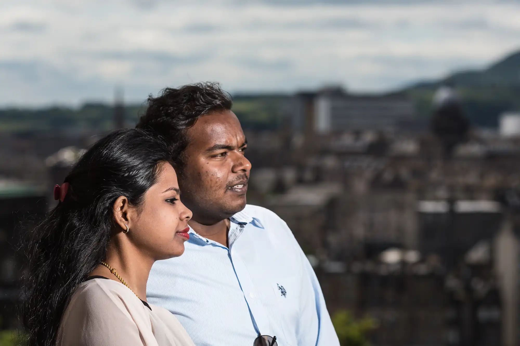 A man and woman standing side by side outdoors, looking into the distance, with a cityscape and hills in the background.