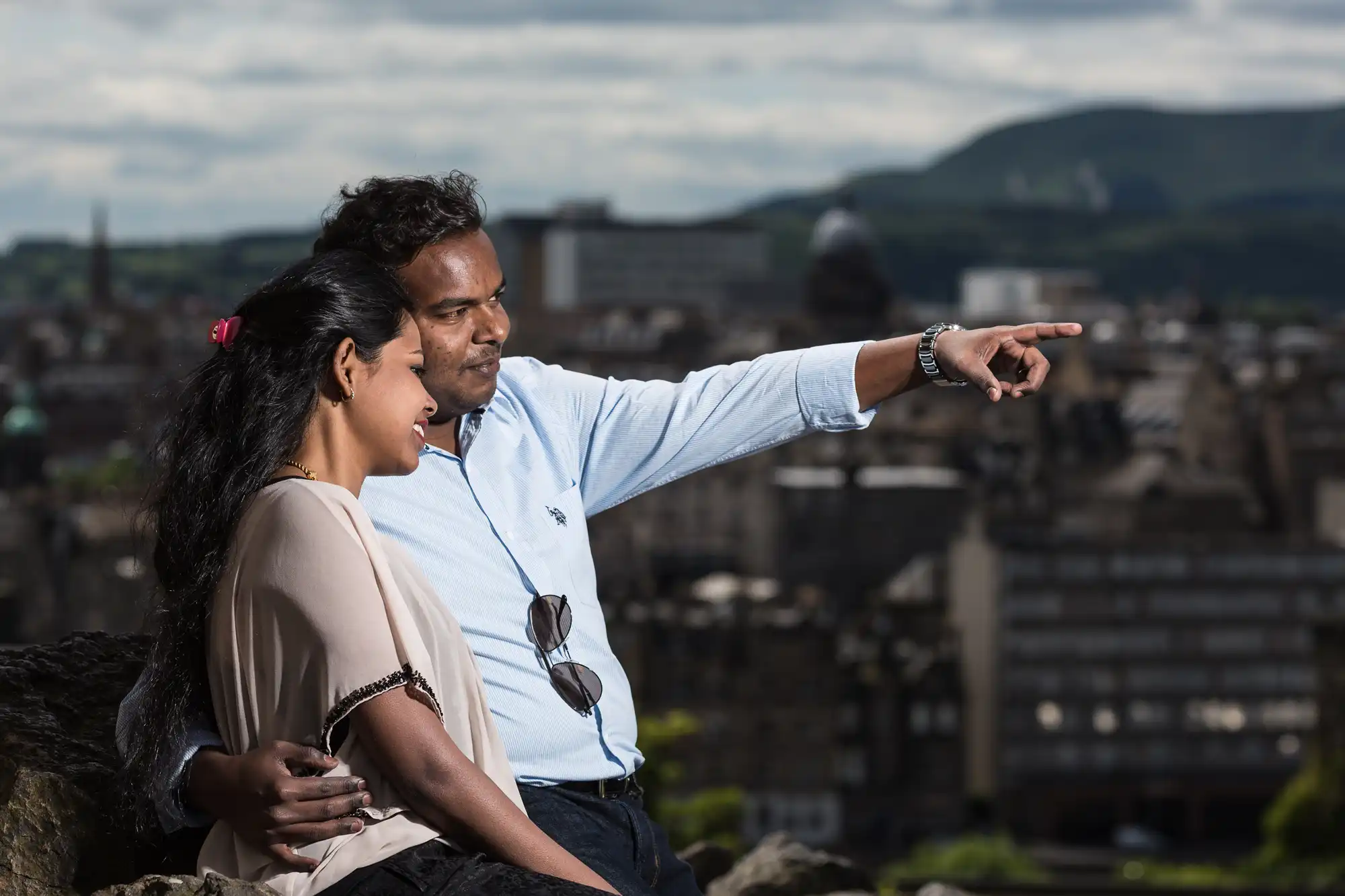 A man and woman sit closely together outdoors with a cityscape in the background. The man is pointing into the distance.