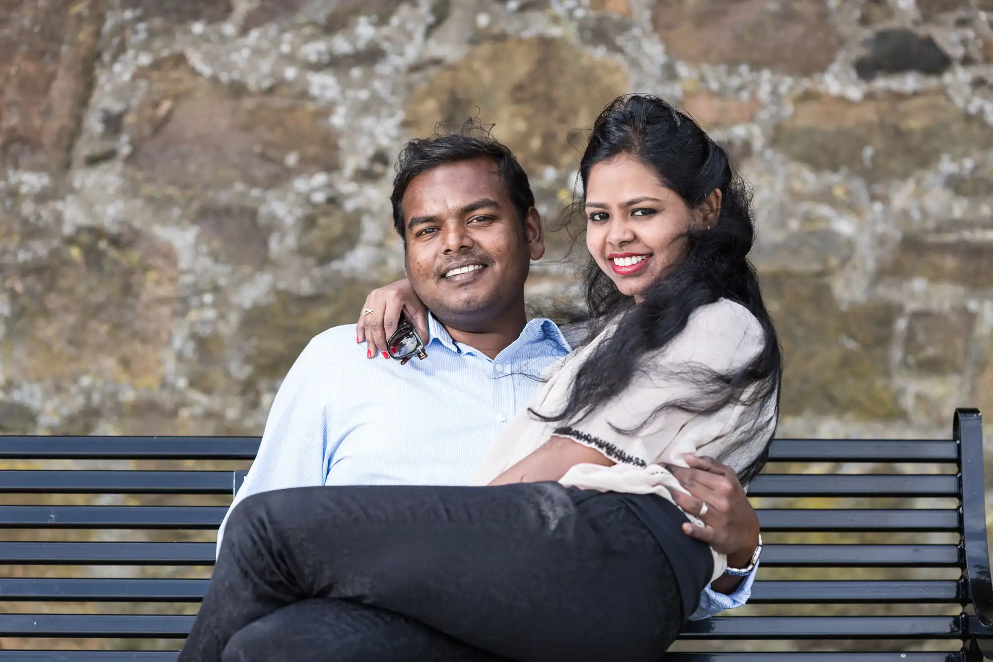 A couple sitting on a bench, smiling at the camera. The woman is partially sitting on the man's lap with her arm around his shoulder. They are in front of a stone wall.