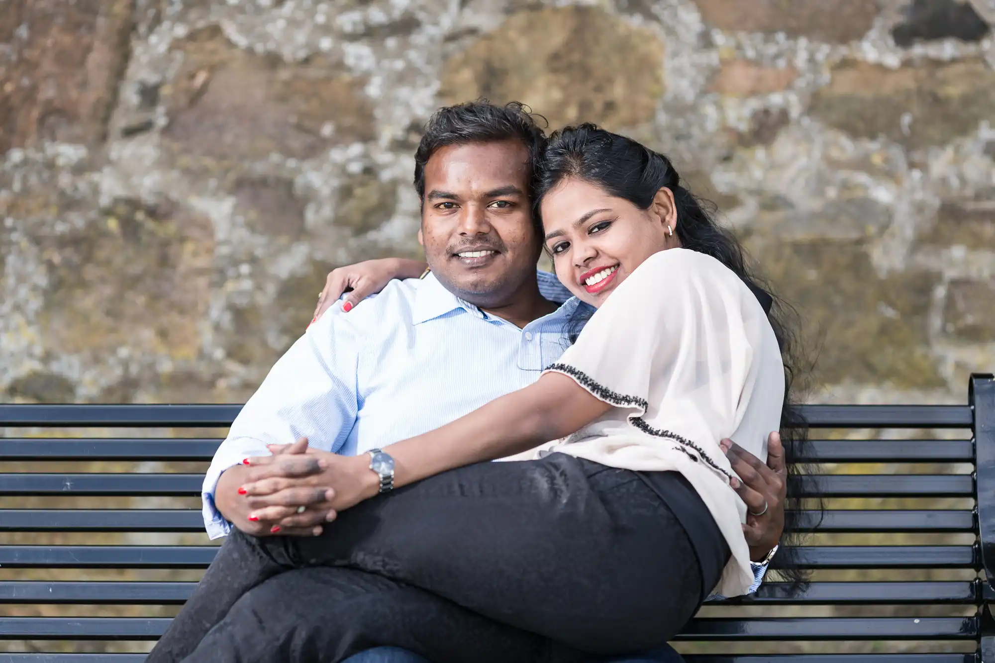 A couple sitting on a bench outdoors; the woman is leaning back into the man's arms, both smiling. Stone wall in the background.