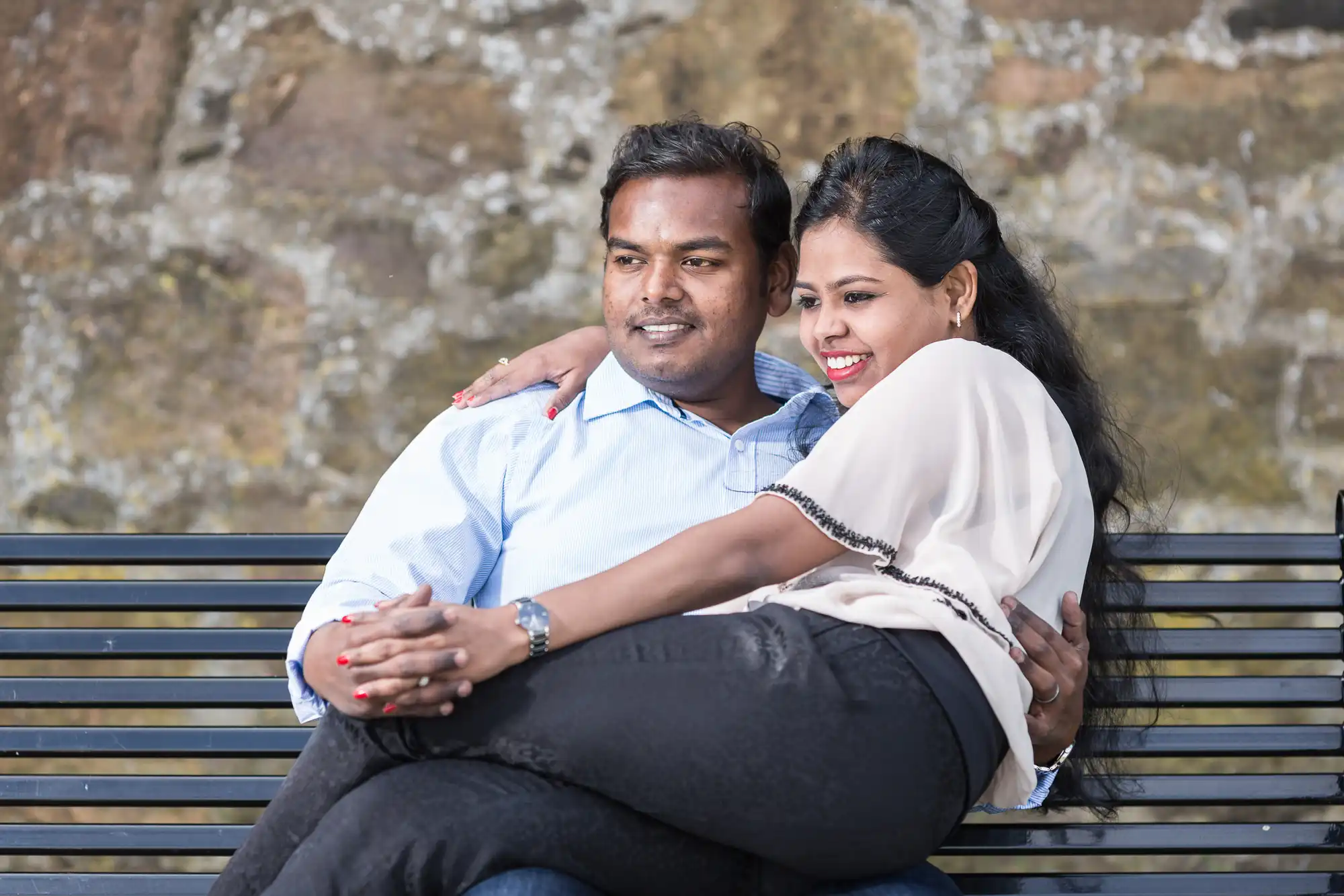 A woman sits on a man's lap on a park bench. Both are smiling and looking ahead. A stone wall serves as the background.