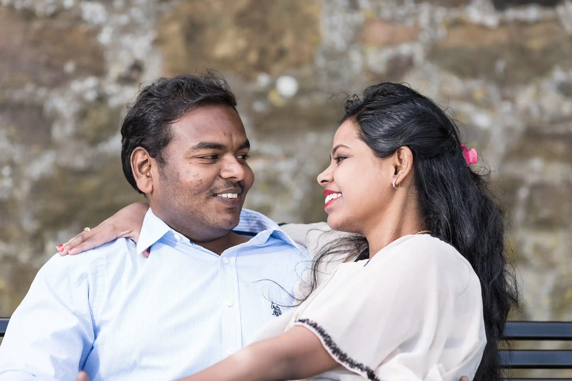 A man and a woman sit closely on a bench outdoors, smiling at each other and engaged in a conversation.