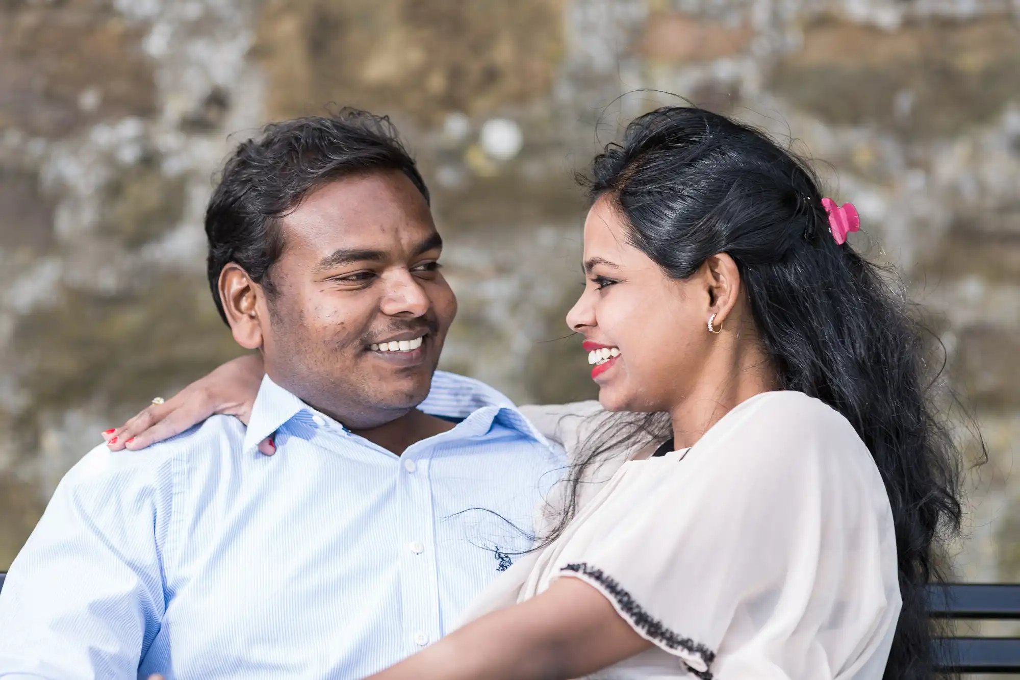 A man and woman are sitting on a bench outdoors. They are smiling at each other.