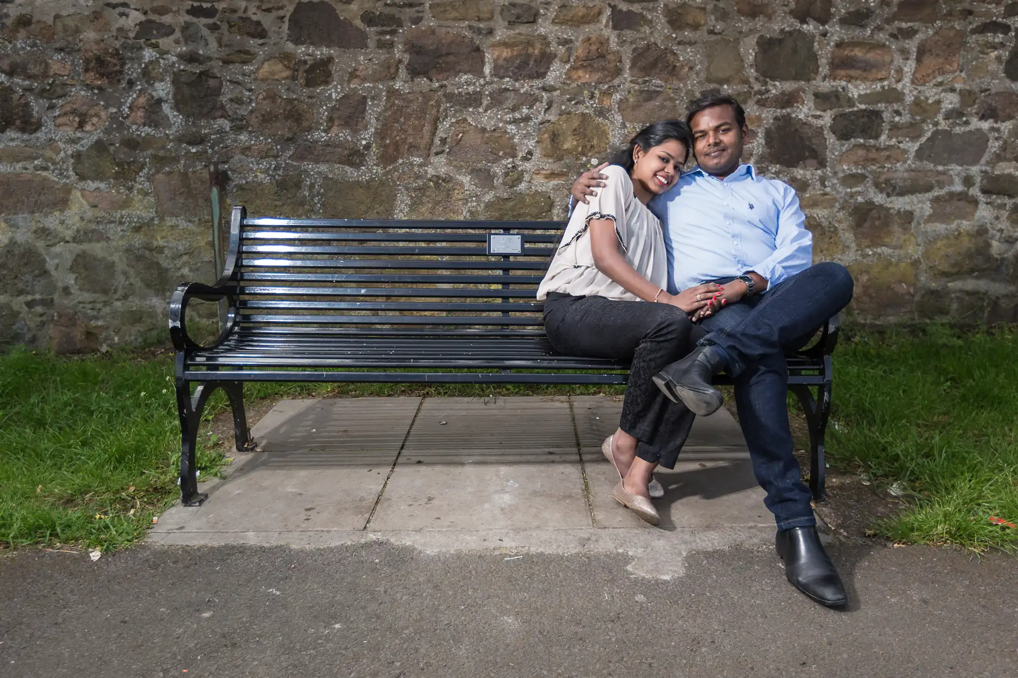 A couple sits on a park bench in front of a stone wall. The woman leans on the man, both smiling, with arms around each other. The setting is outdoors with grass and pavement around the bench.