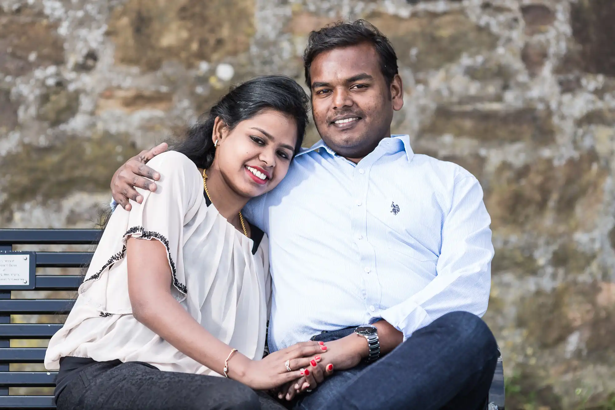 A couple sits closely on a bench, smiling and embracing each other. The background features a textured stone wall. Both are dressed in casual clothing.