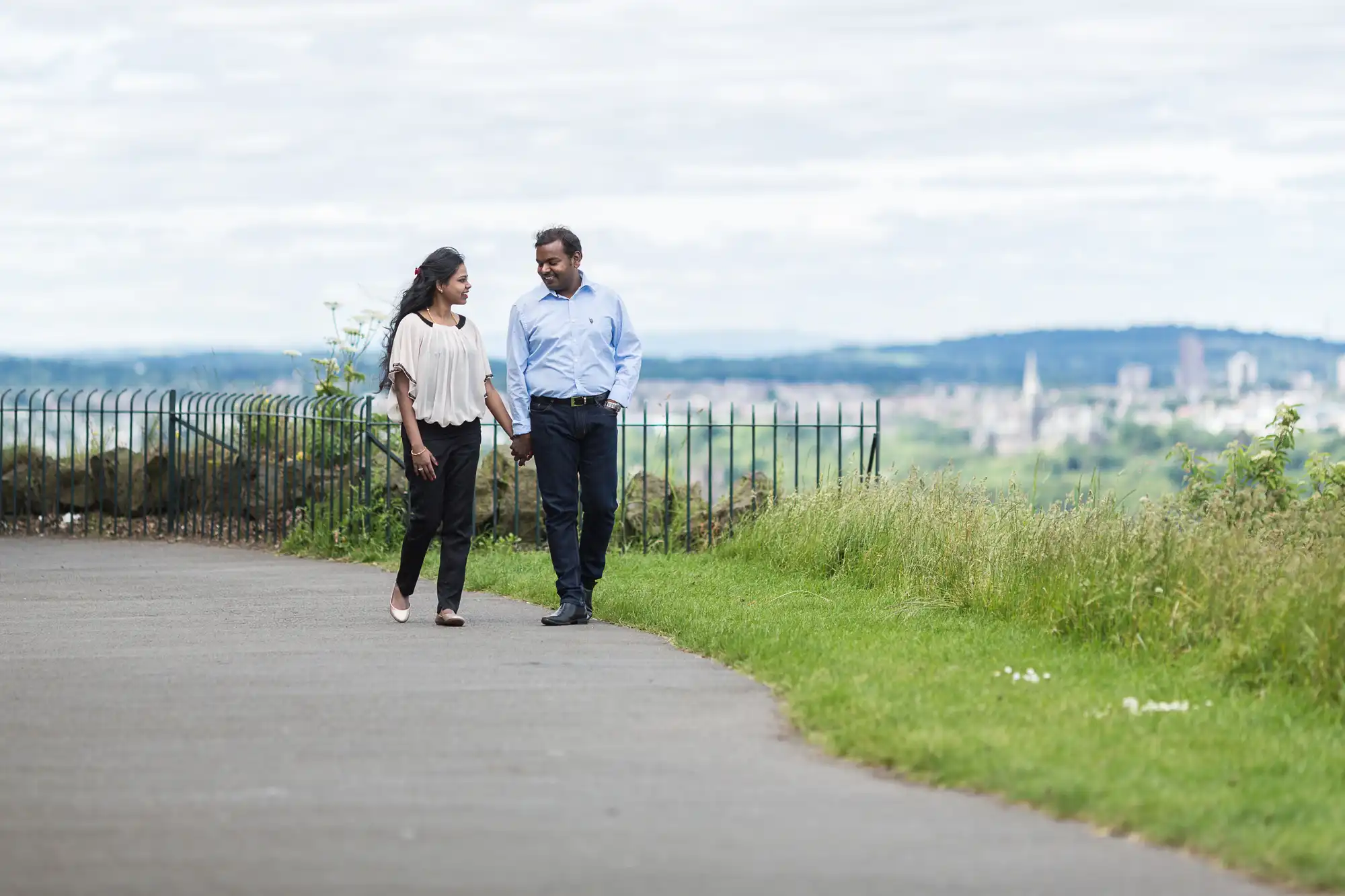 A man and woman walk hand-in-hand along a paved path lined with grass and a metal fence, with a cityscape and cloudy sky in the background.